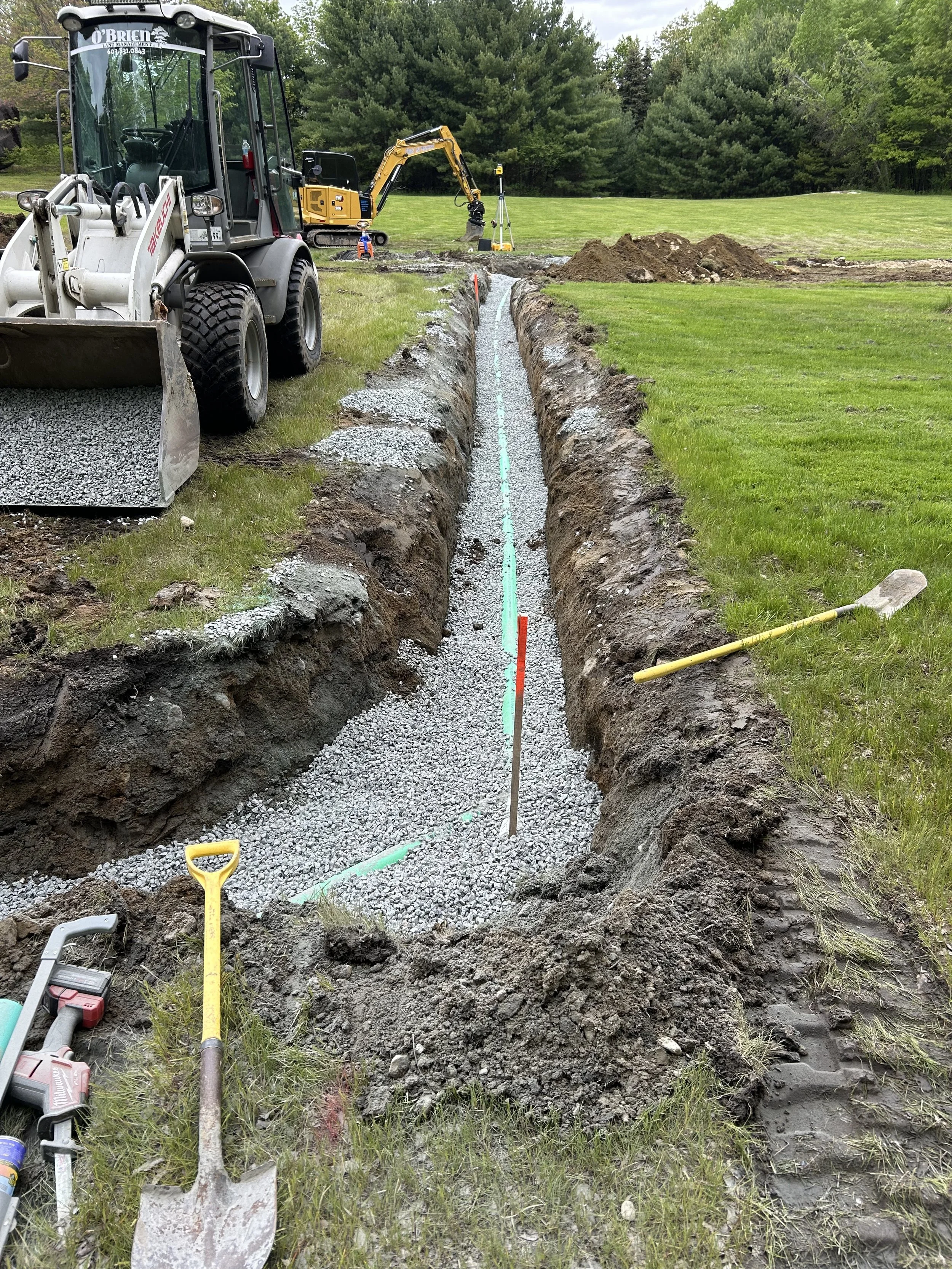 Construction site featuring a trench for underground utility installation with construction tools, machinery, and equipment in a grassy field with trees in the background.