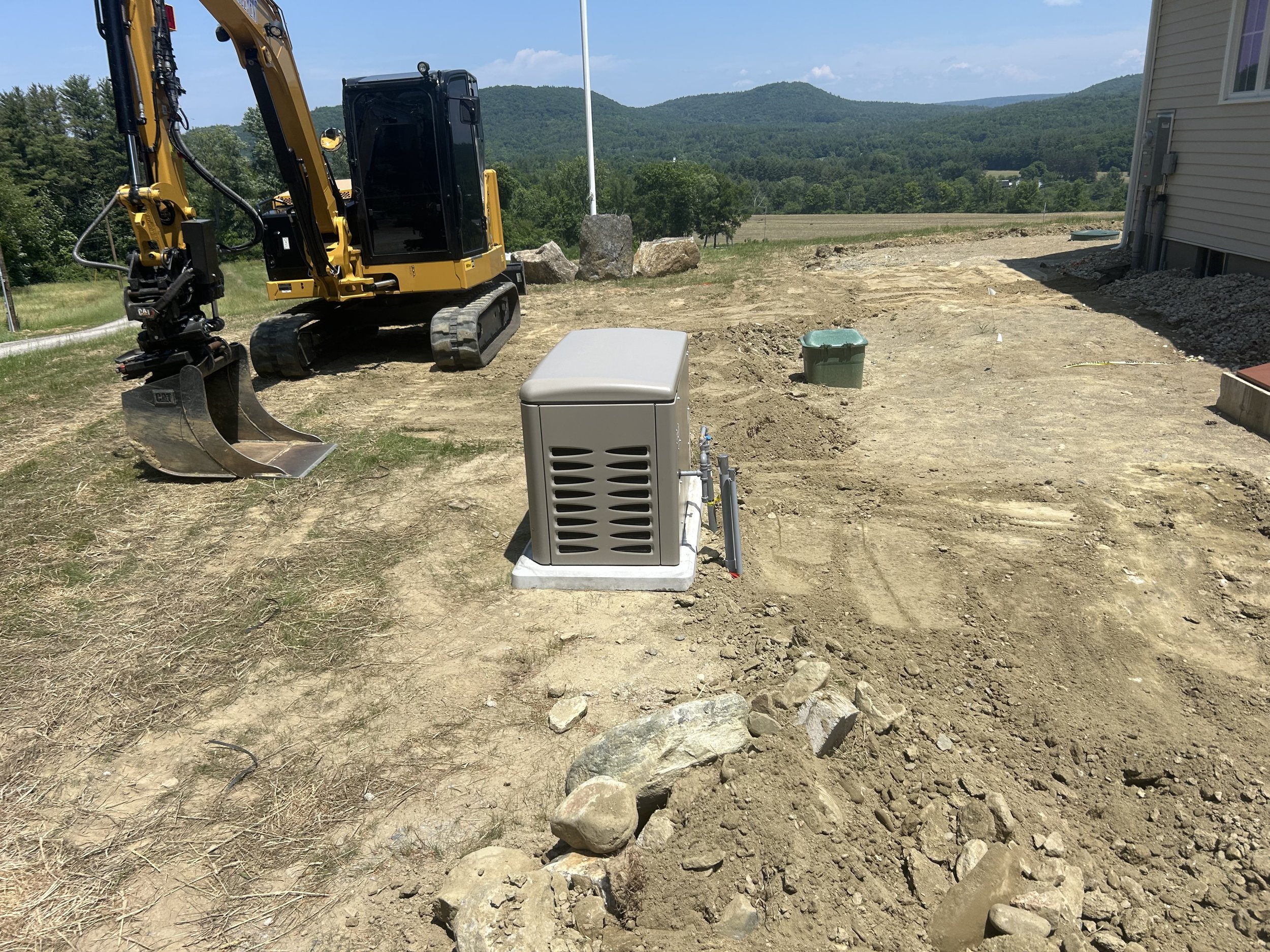 Construction site with a small excavator, a utility box, a green container, and a dirt pathway, with a hilly landscape in the background.