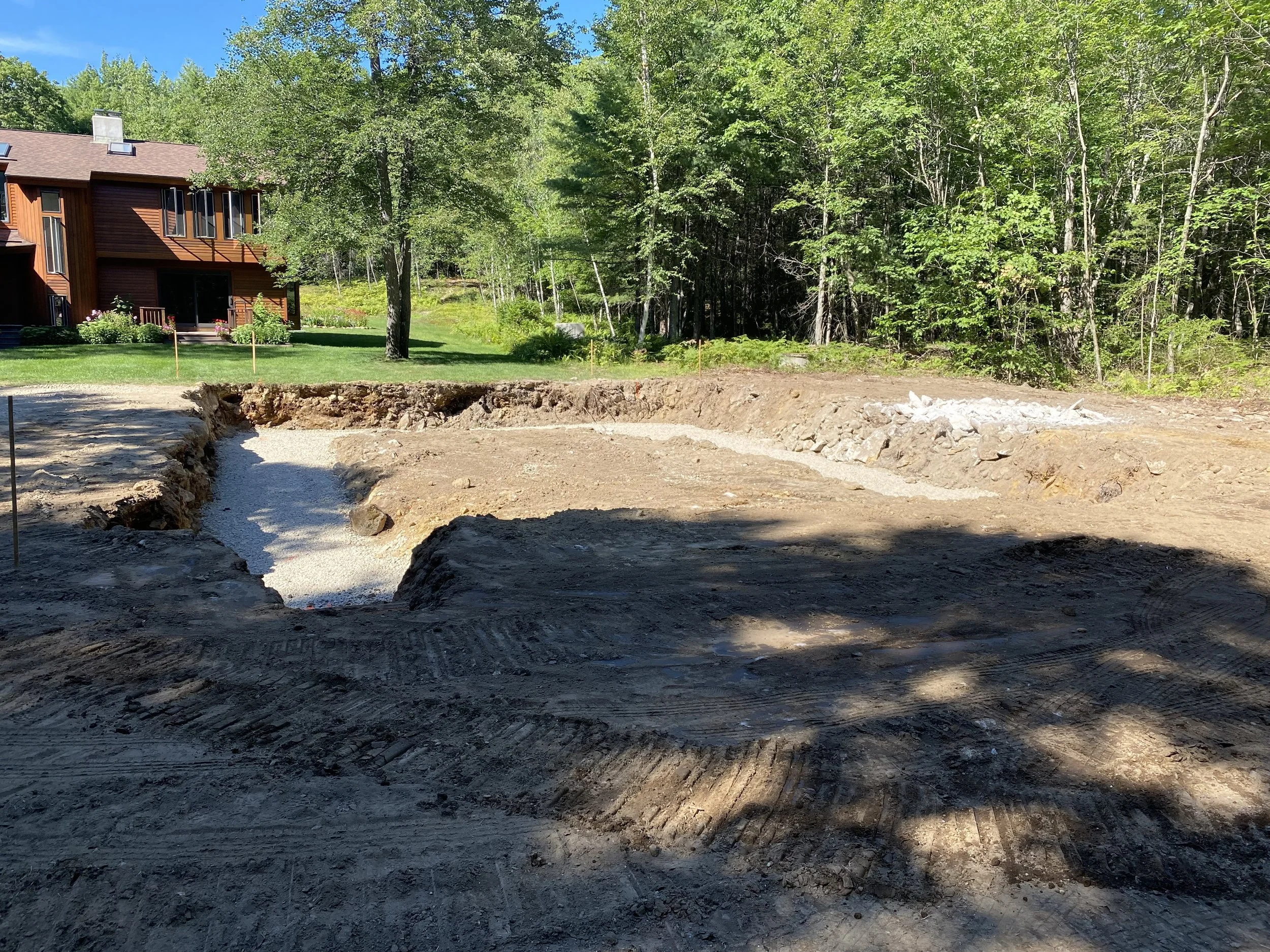 A construction site with a cleared and excavated area, surrounded by trees and a house in the background.
