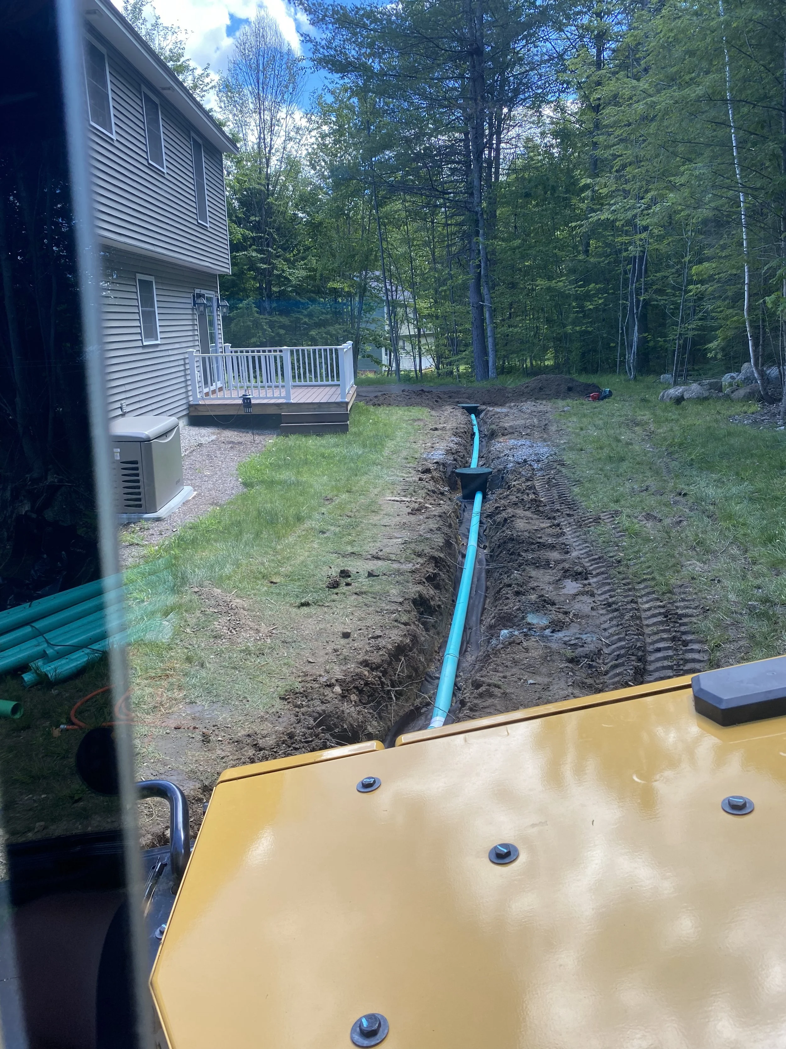 A construction site showing a trench dug alongside a house, with blue piping installed in the trench and black pipe fittings attached. The scene is outdoors with a house on the left and a wooded area in the background.