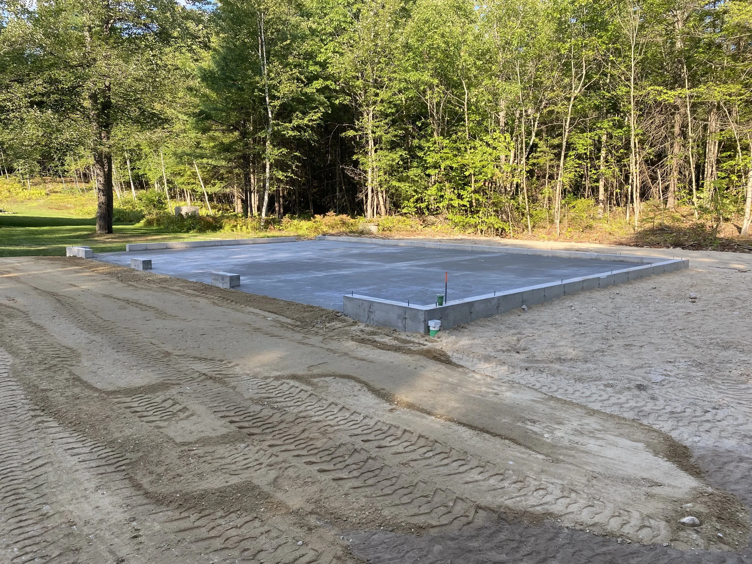 Construction site of a building foundation with concrete slab and concrete block walls, surrounded by dirt and tire tracks, with trees and greenery in the background.
