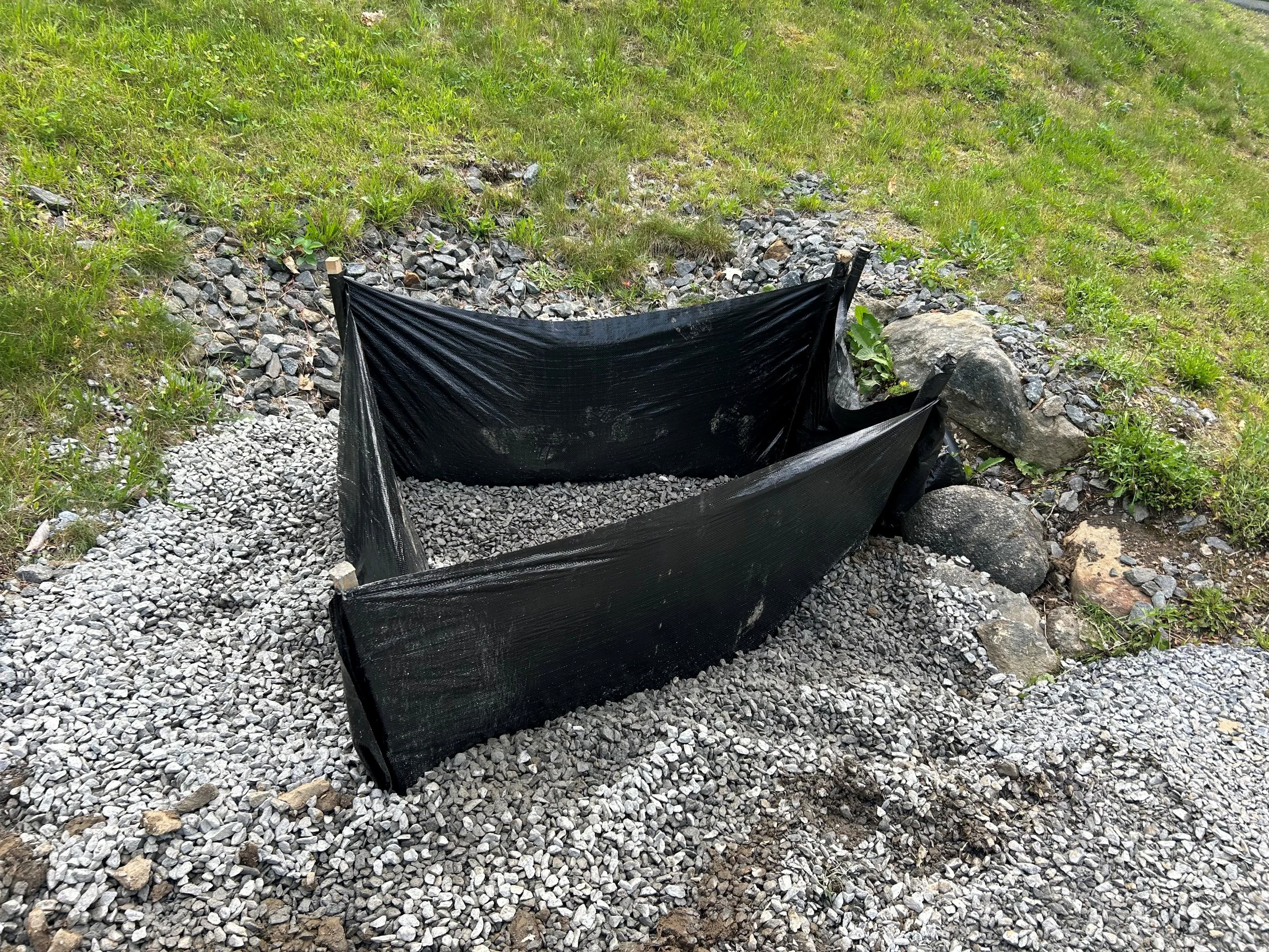 A small, makeshift garden bed or pond enclosure made from black plastic sheeting and rocks, set on a gravel surface, surrounded by grass and rocks.