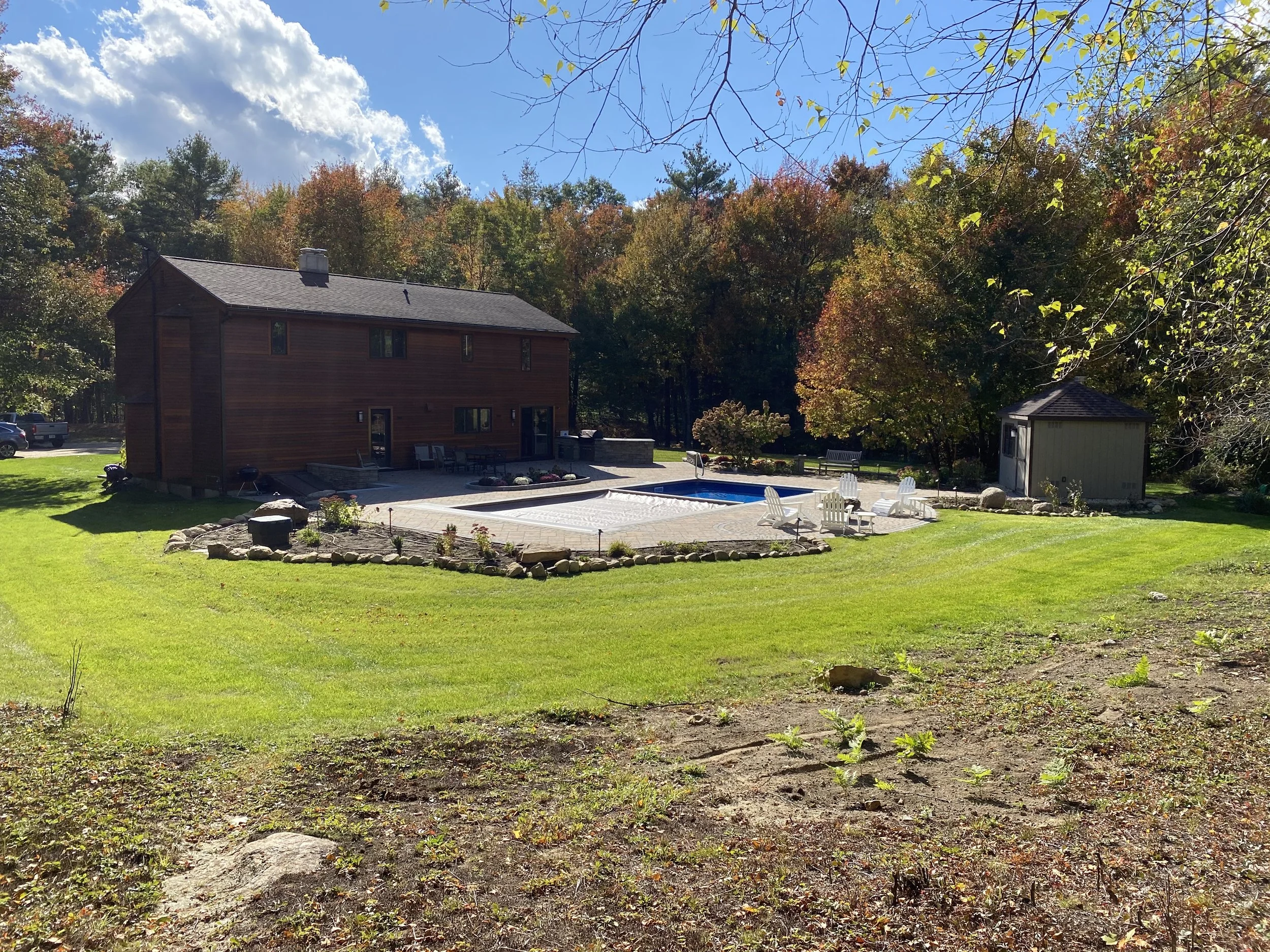 A backyard with a patio, swimming pool, Adirondack chairs, a small shed, and a wooden house, surrounded by trees with fall foliage.