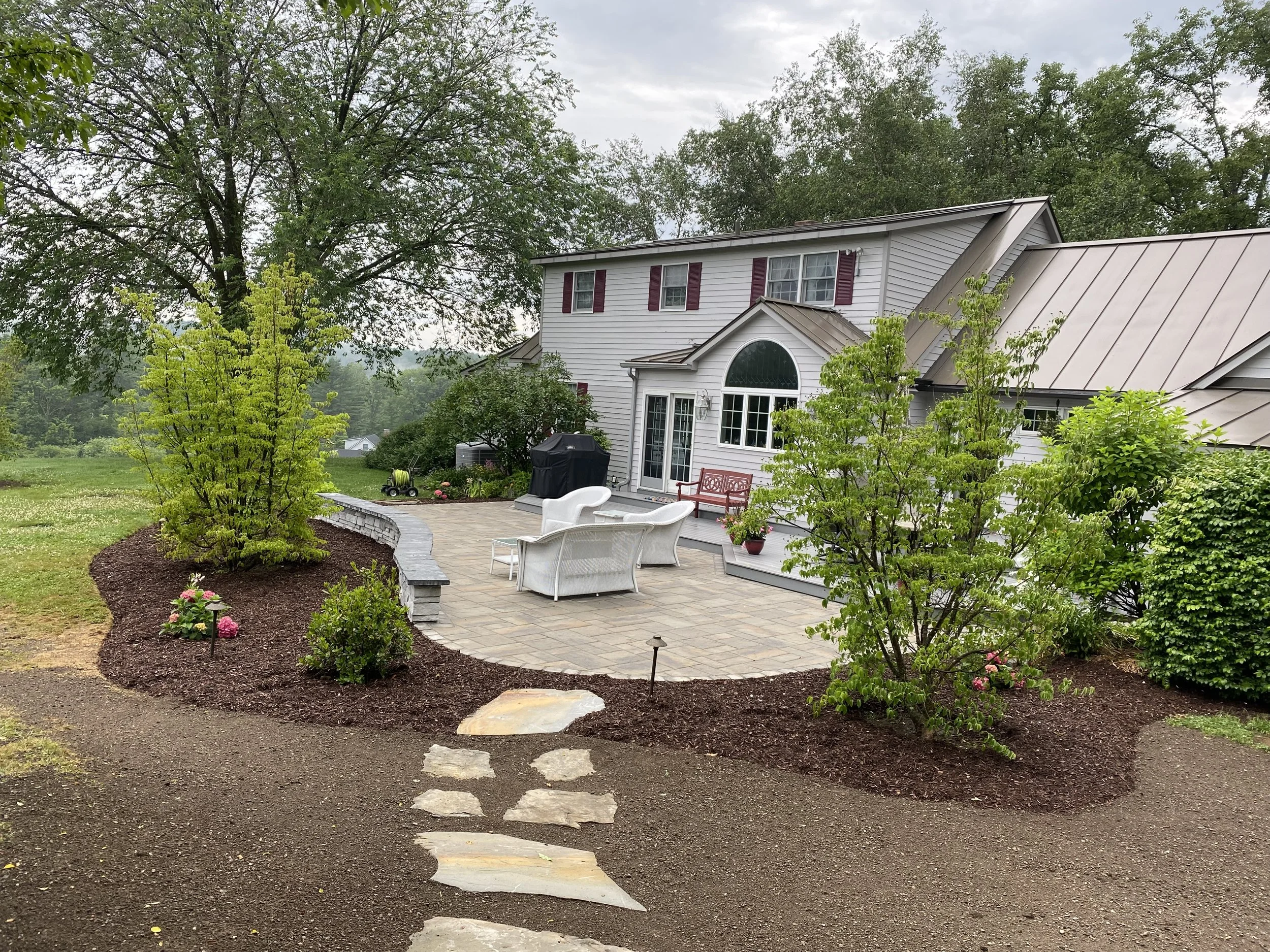 Backyard patio area with outdoor furniture, potted plants, and a barbecue grill, surrounded by landscaped flower beds and a large tree, with a house in the background.