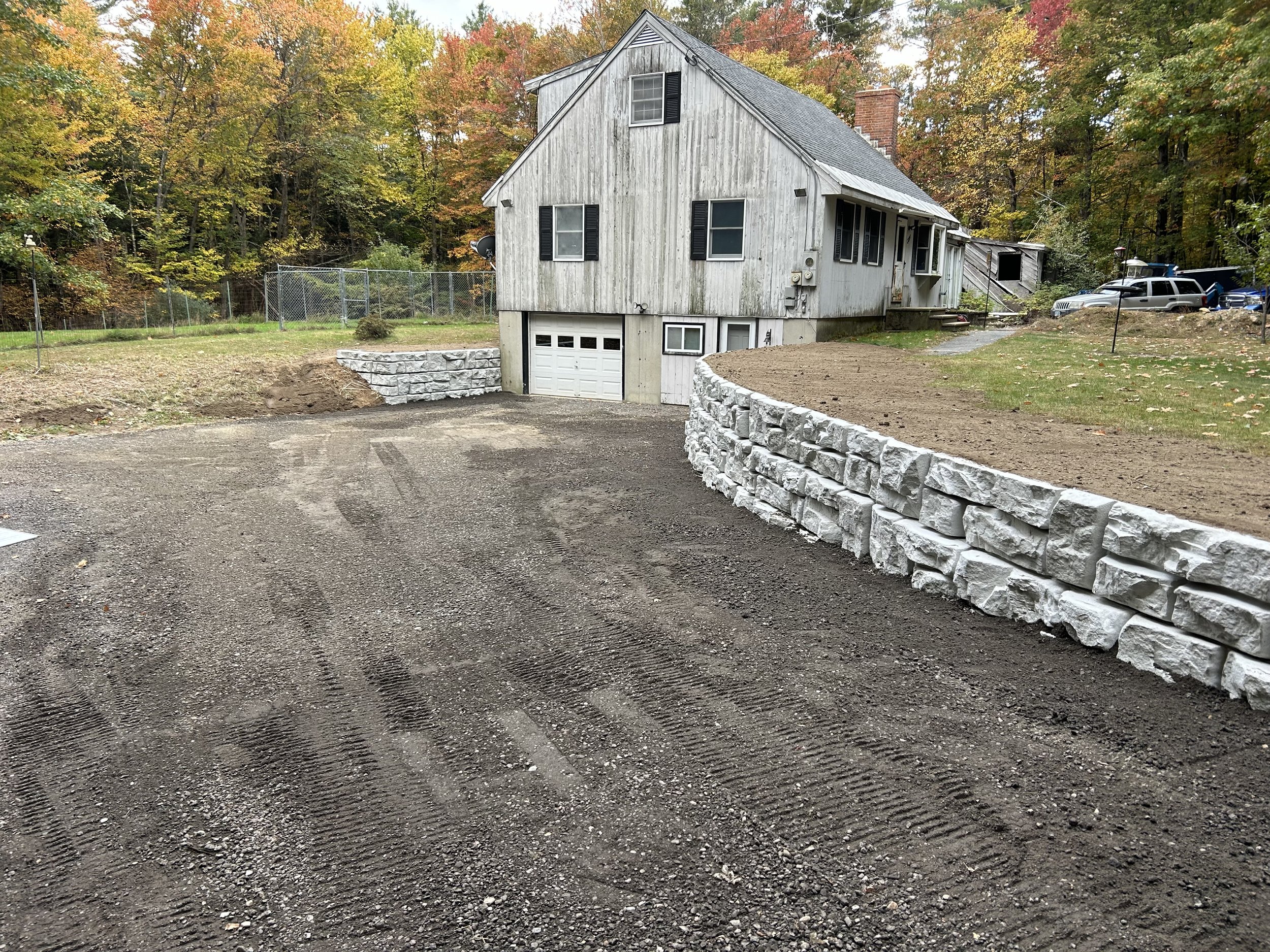 A house with a garage built into a sloped hill, surrounded by a newly constructed white stone retaining wall and a dirt driveway, with a wooded background of autumn trees.