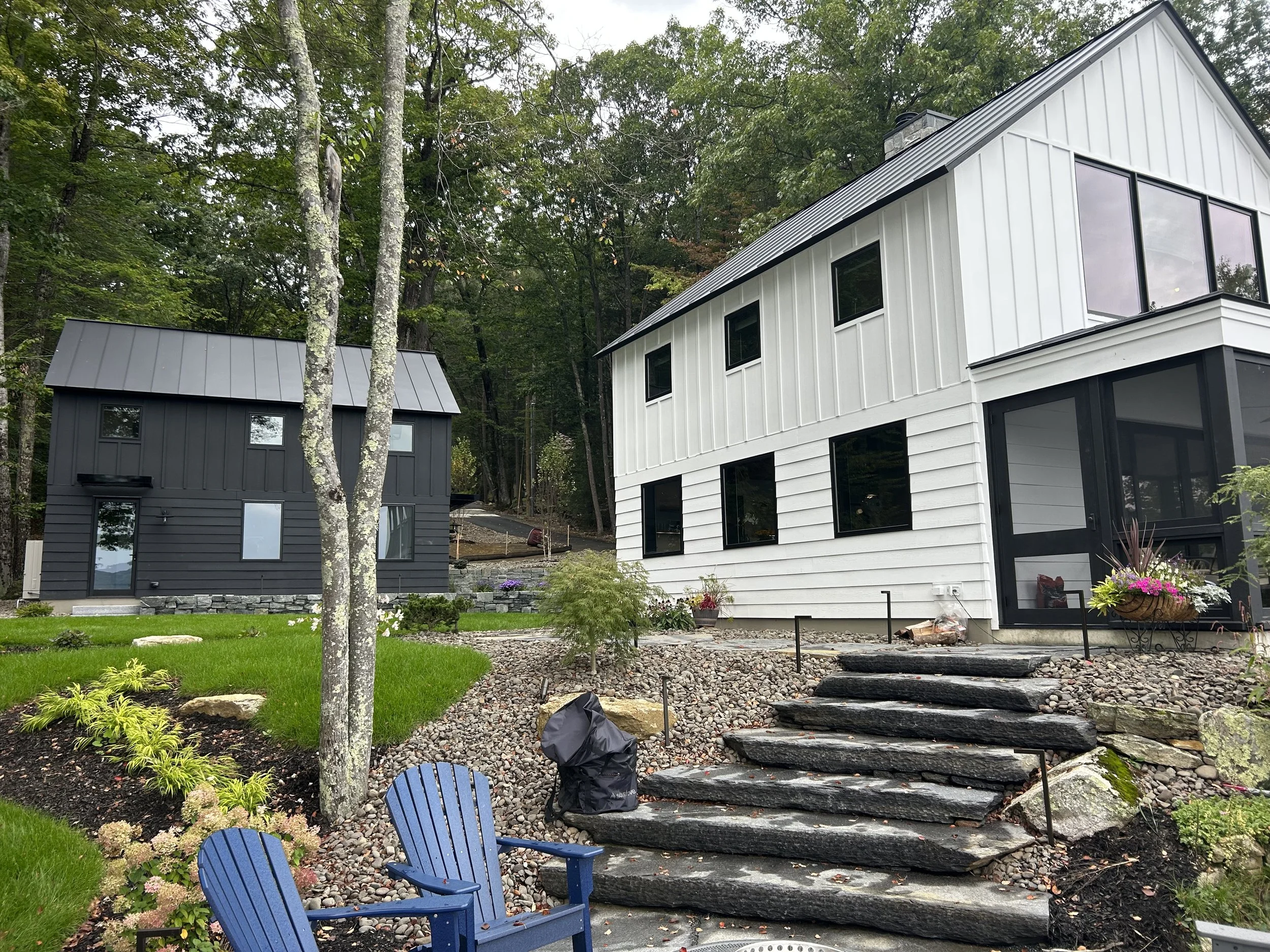 Two modern houses, one painted black and the other white, are located in a landscaped yard with stone steps, greenery, and trees in the background.
