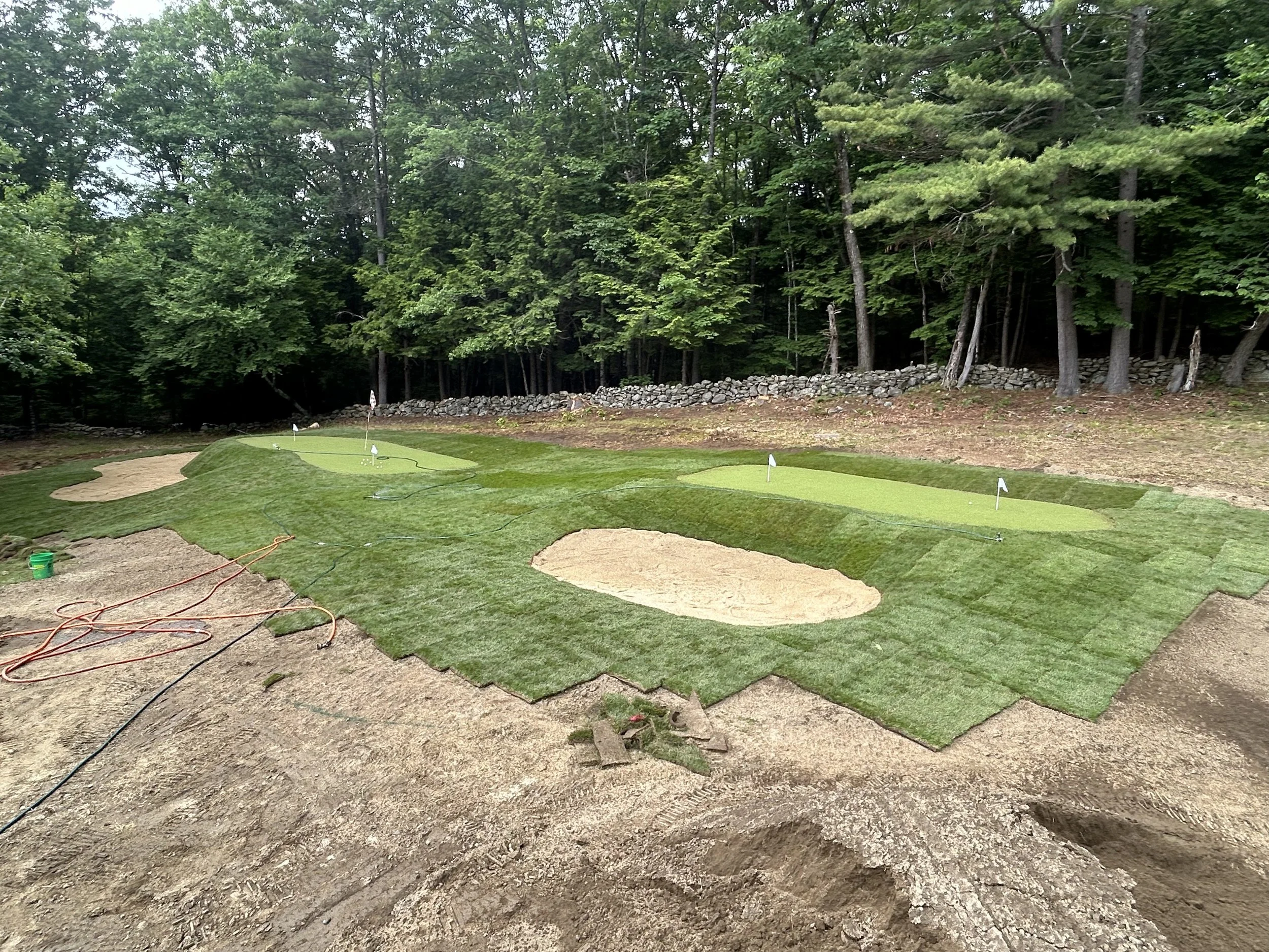 A golf practice area with two putting greens, sand bunkers, and grass patches, surrounded by trees and a stone wall in the background.