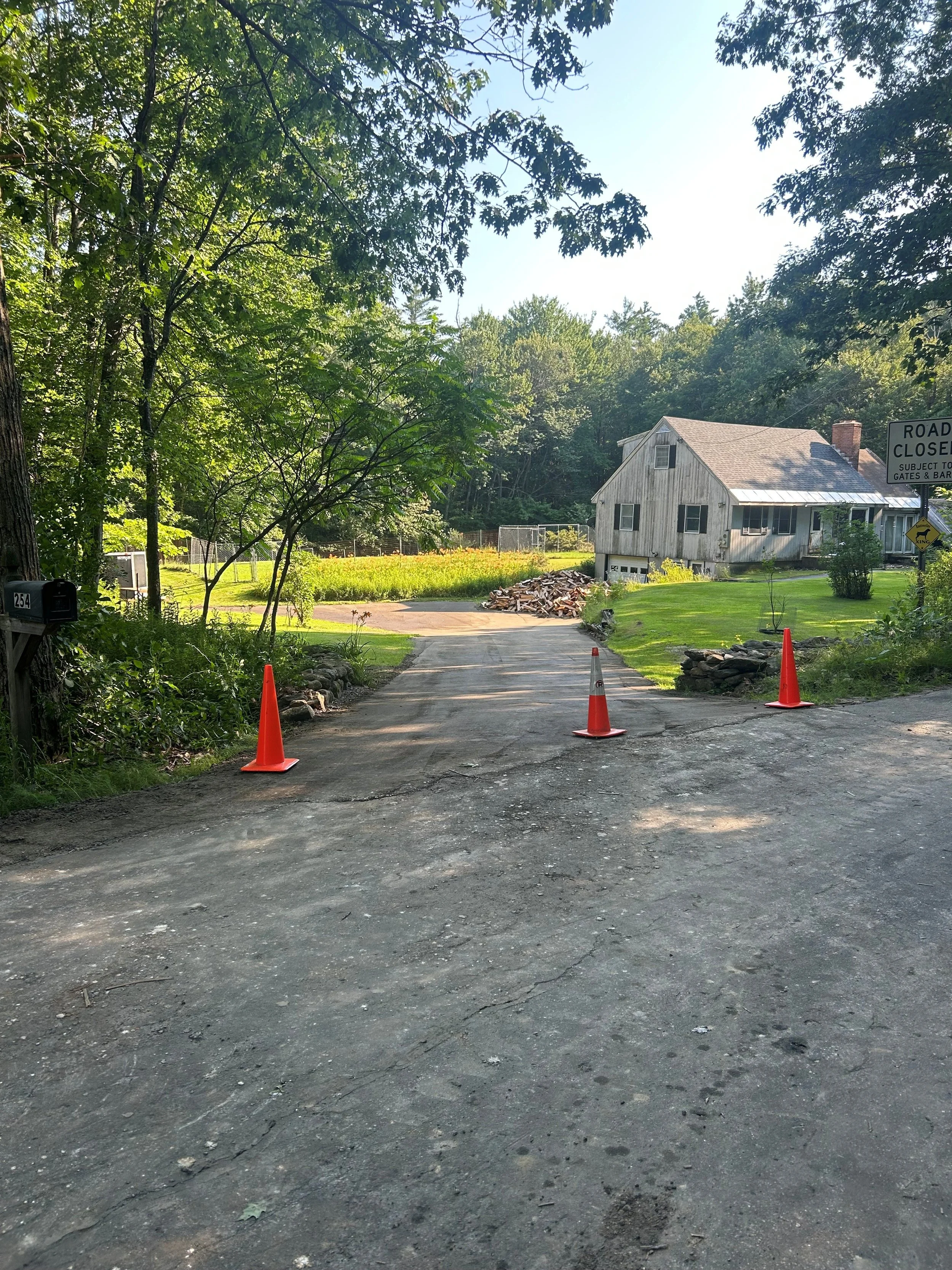A house with a yard behind a dirt and asphalt road blocked by three orange traffic cones, with a sign indicating the road is closed, and a stack of firewood on the driveway.