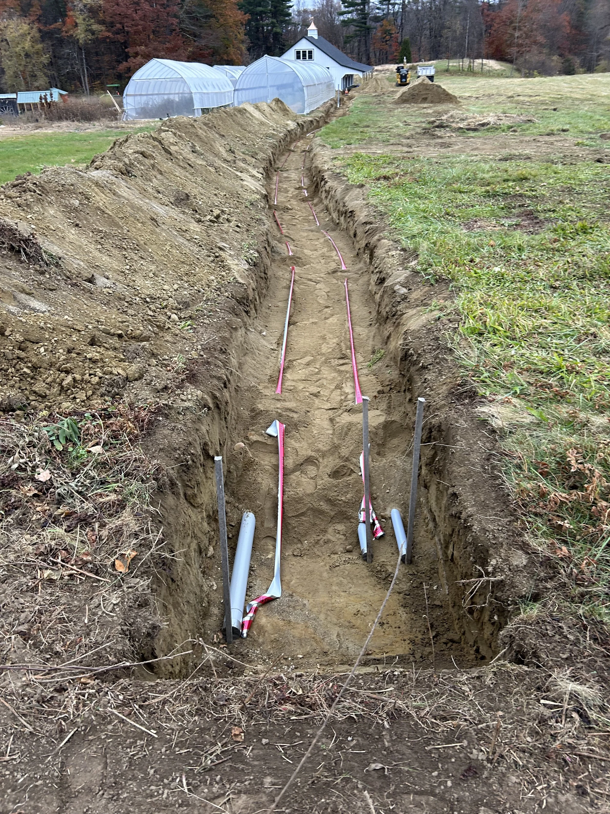 A construction trench with exposed pipes, located on a grassy field with a greenhouse and a white barn in the background.
