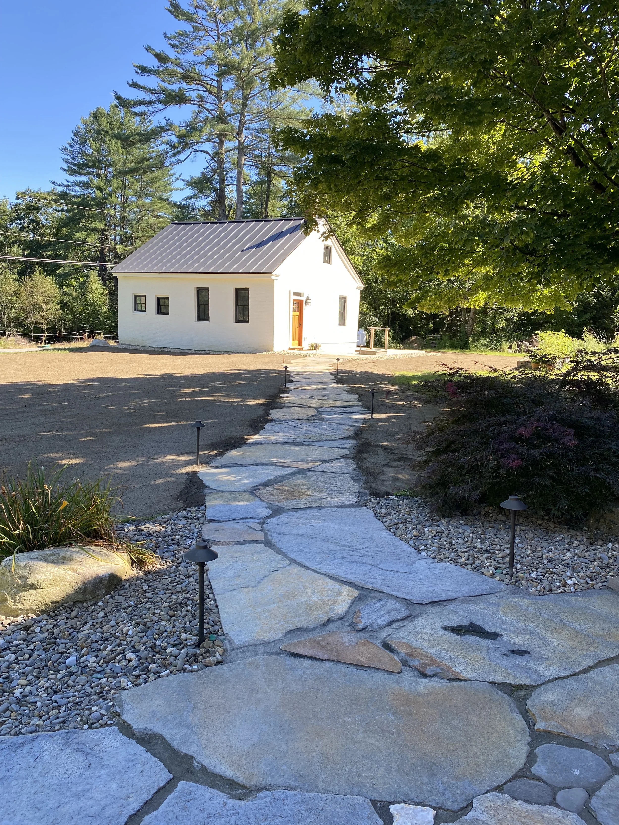 A white house with a metal roof, situated at the end of a stone pathway, surrounded by greenery, trees, and a clear blue sky.