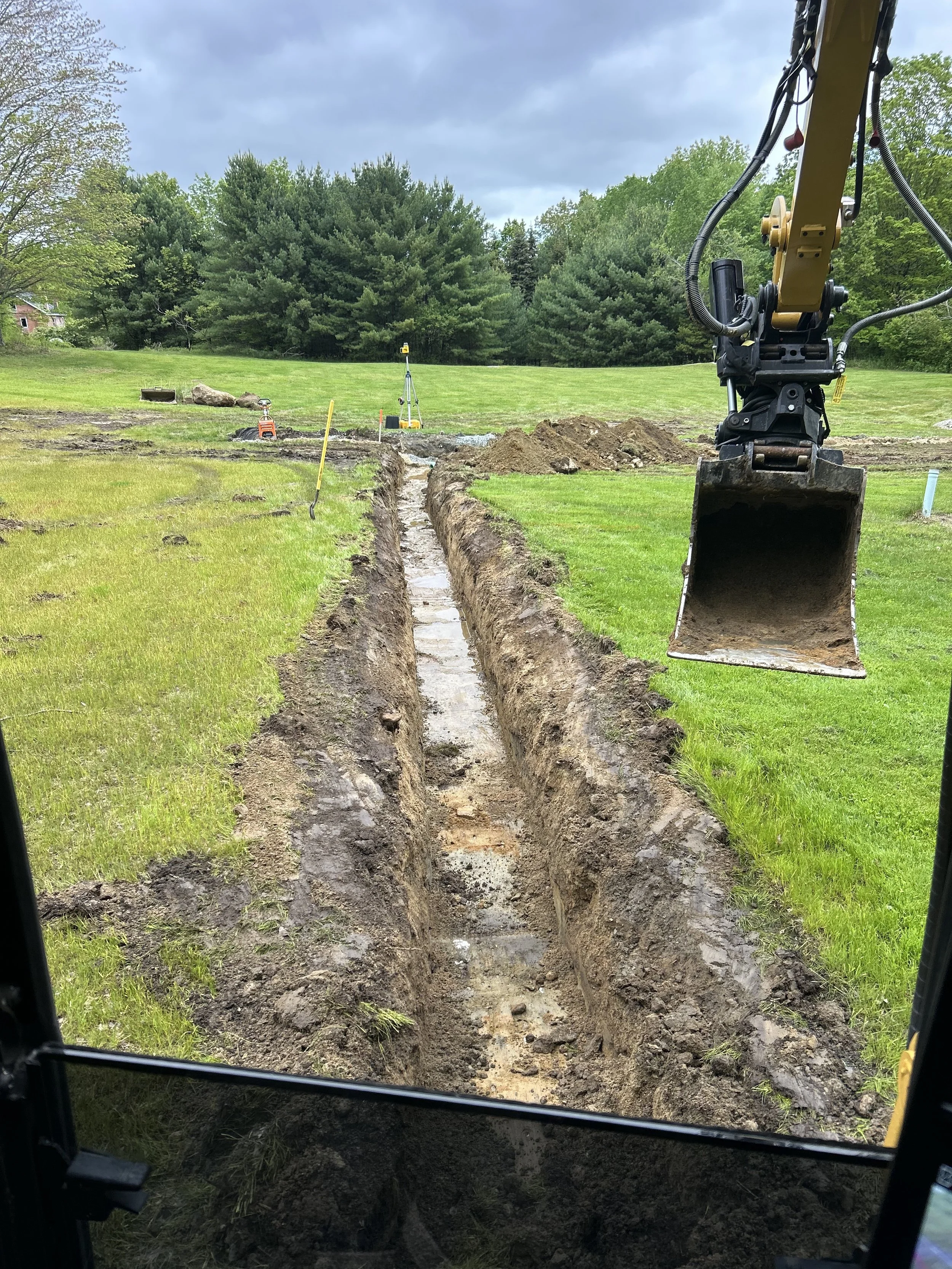 View from inside a construction vehicle looking out over a trench being excavated in a grassy yard, with trees in the background and construction tools and equipment at the far end.
