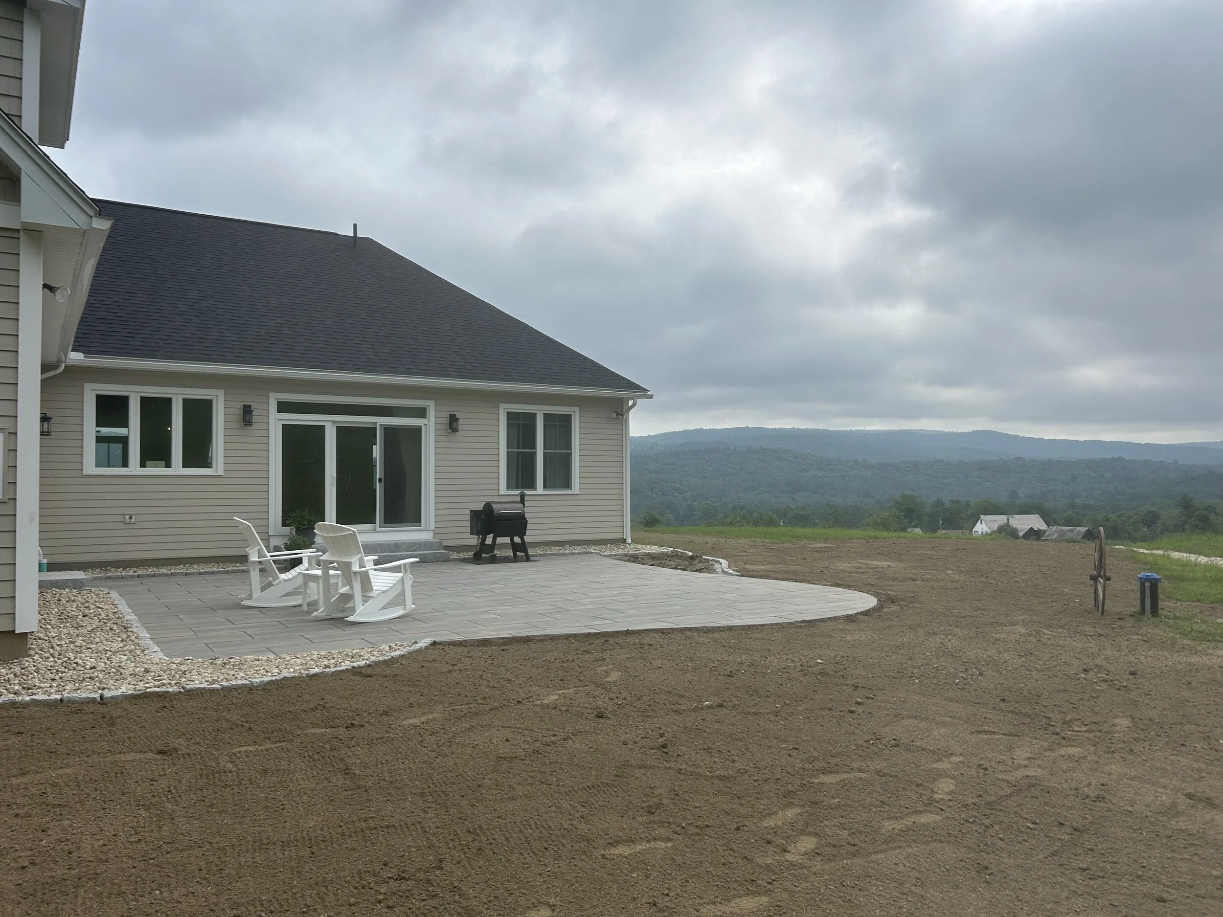 View of a house with a patio, white chairs, a grill, and a rural landscape with hills and overcast sky.