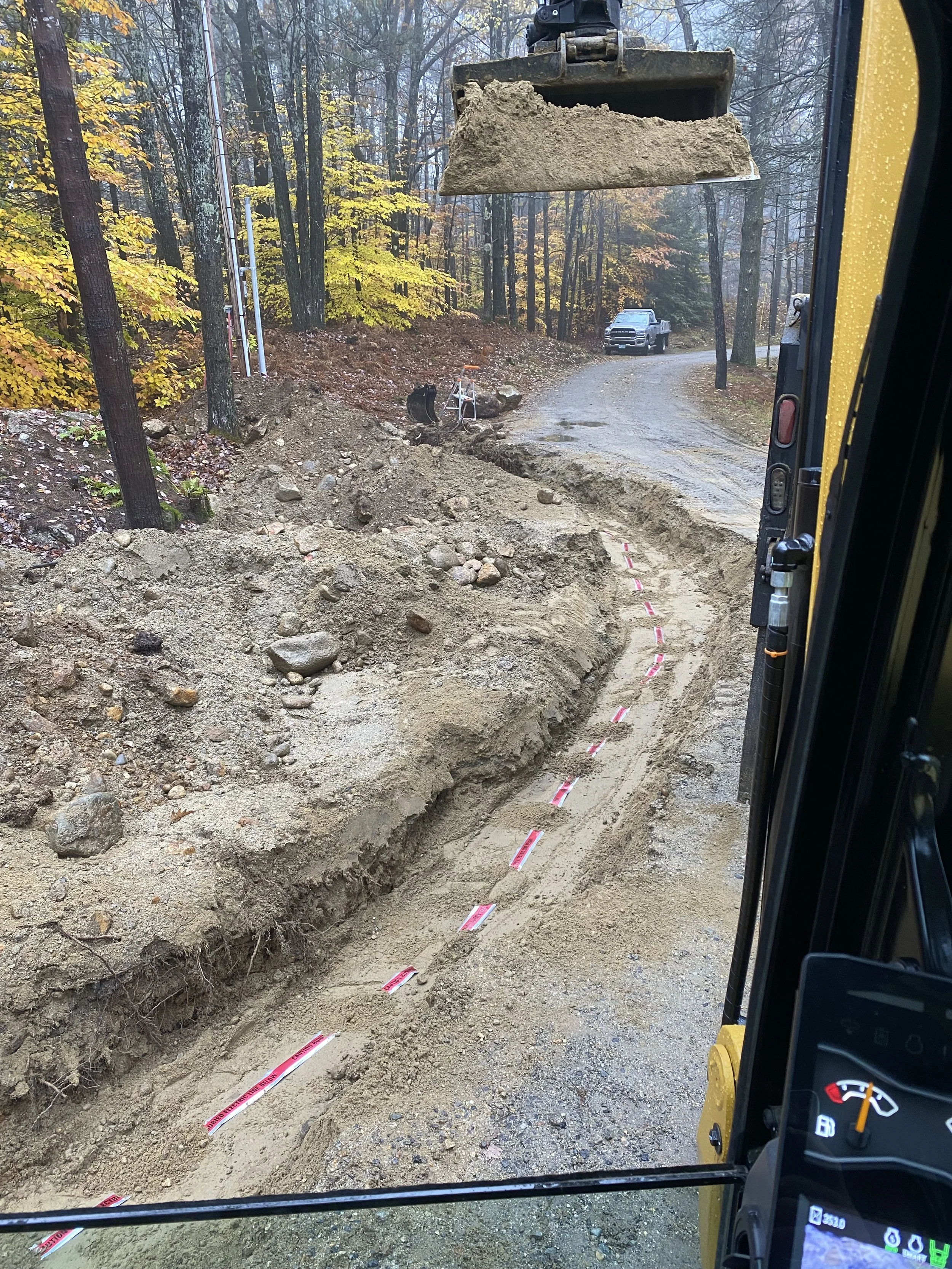 Construction site on a dirt road through a forest, with a backhoe digging a trench, a damaged section of the road, and a pickup truck parked further along the road. Forest trees with fall foliage surround the scene.