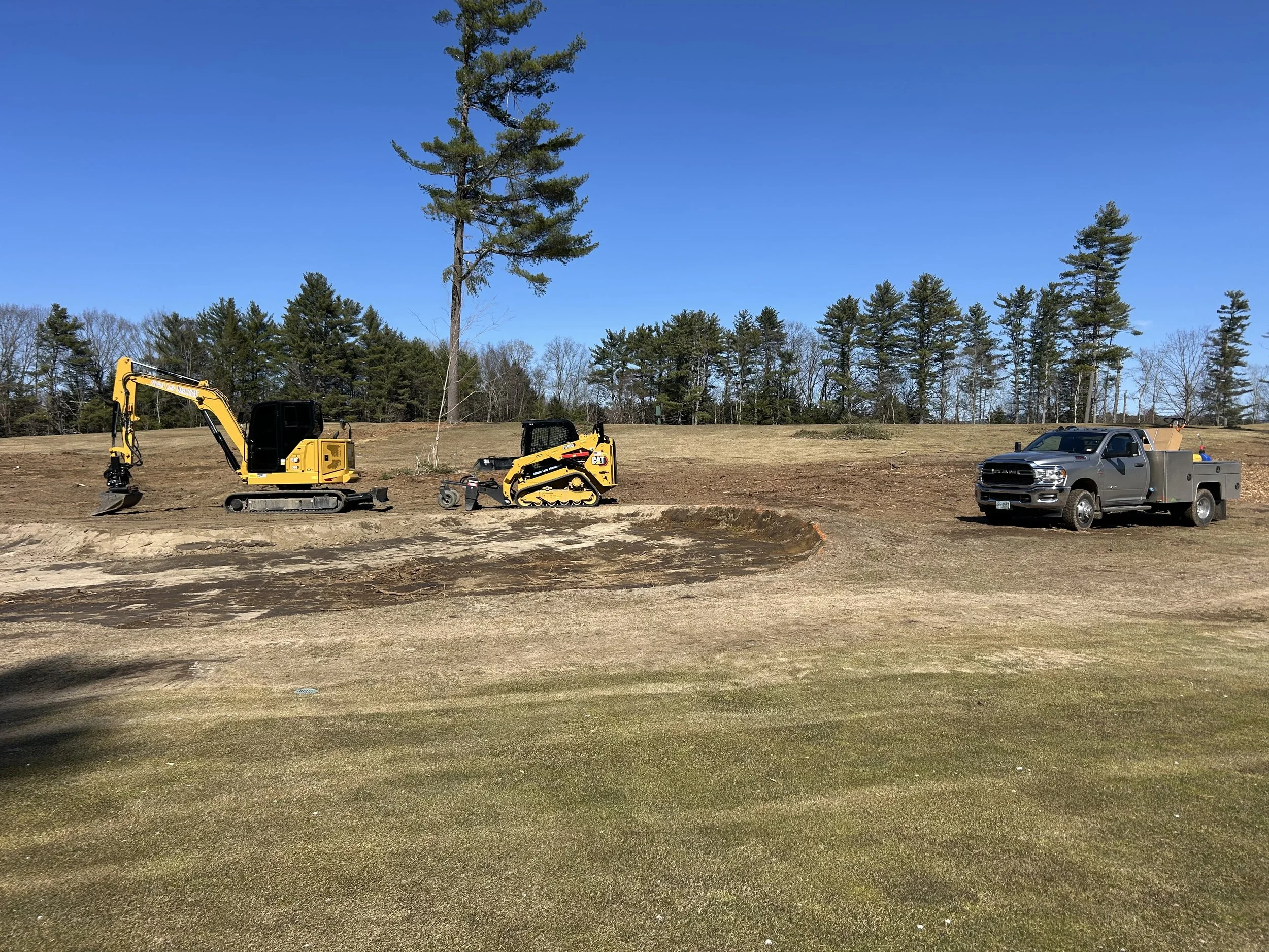 A construction site with two yellow mini excavators and a gray work truck parked on a partly grassy hill, with a row of trees and a blue sky in the background.