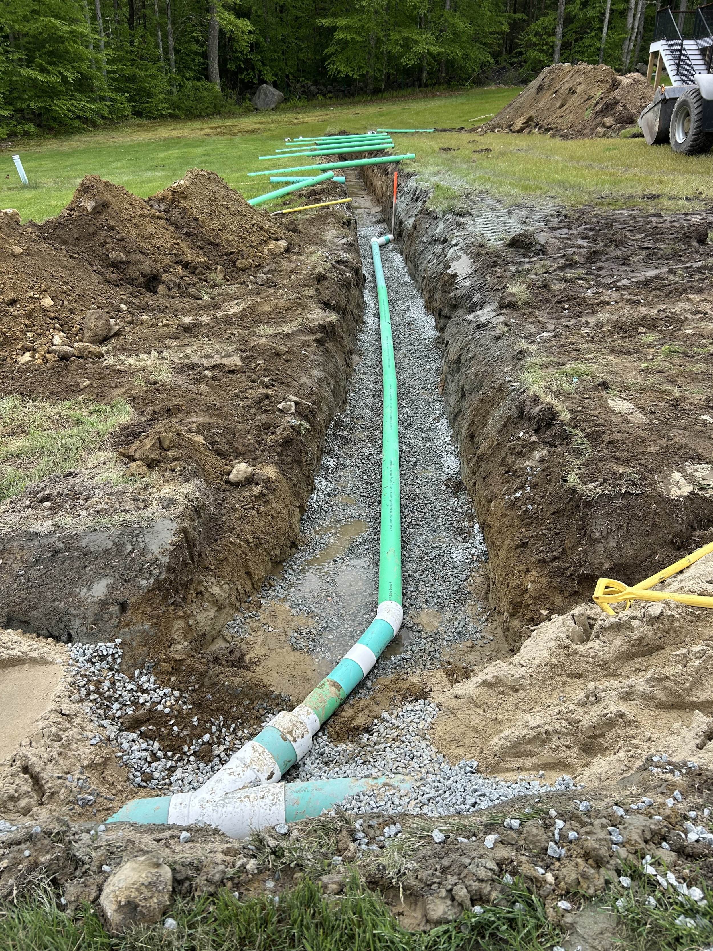 Underground water pipeline installation in progress, with green pipes laid in a trench surrounded by dirt and gravel, outdoors in a grassy area with trees in the background.
