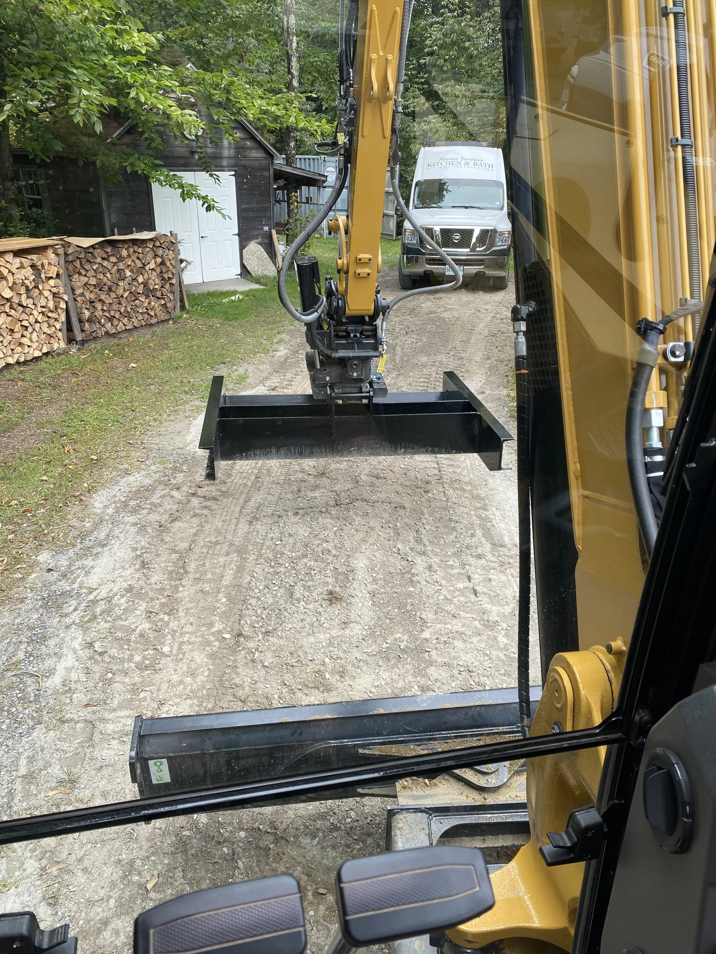 View from inside a construction vehicle looking out over a dirt road with a hydraulic arm and attachment in the foreground, a white utility truck in the background, a small black shed, and stacks of chopped firewood on the left.