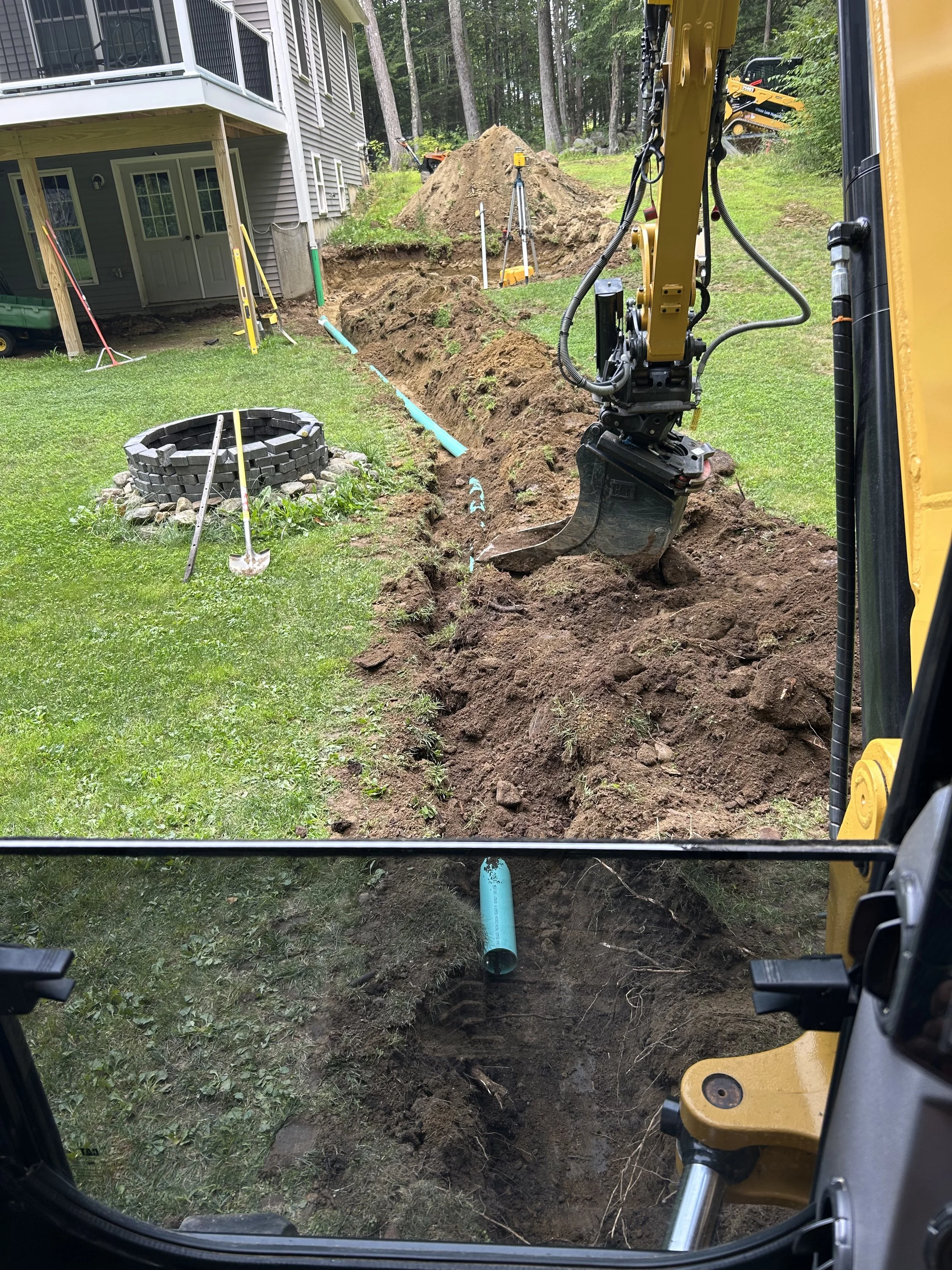 Excavator digging a trench in a backyard for underground piping, with construction tools and a fire pit nearby.