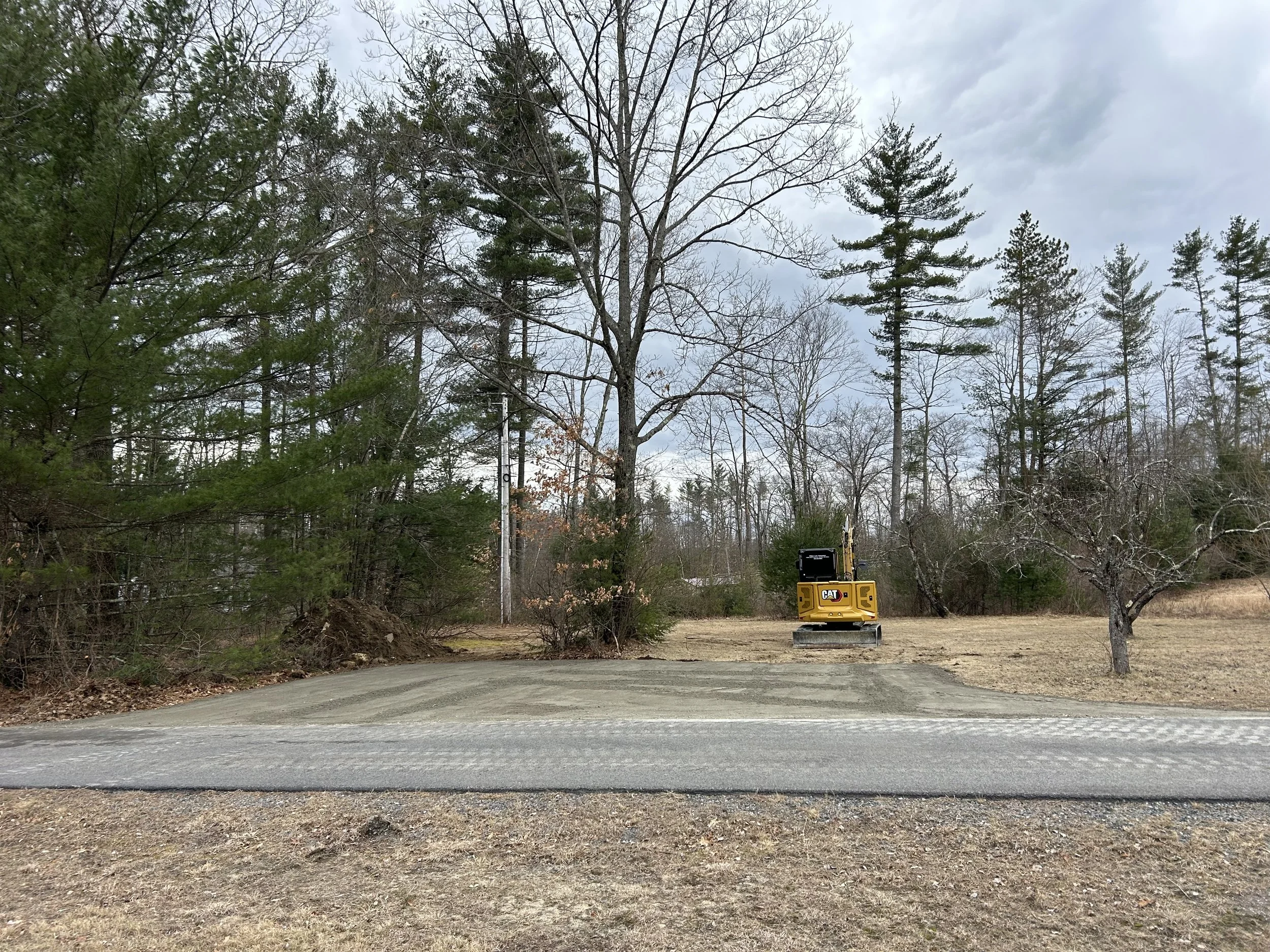 A yellow compact excavator parked on a gravel patch next to a wooded area with trees, some of which are leafless, under a partly cloudy sky.