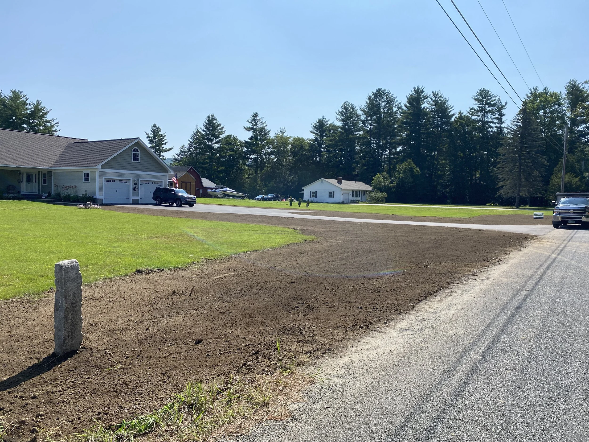 A residential neighborhood with houses, cars parked on driveways and street, green lawns, a patch of freshly tilled dirt, a clear blue sky, and power lines