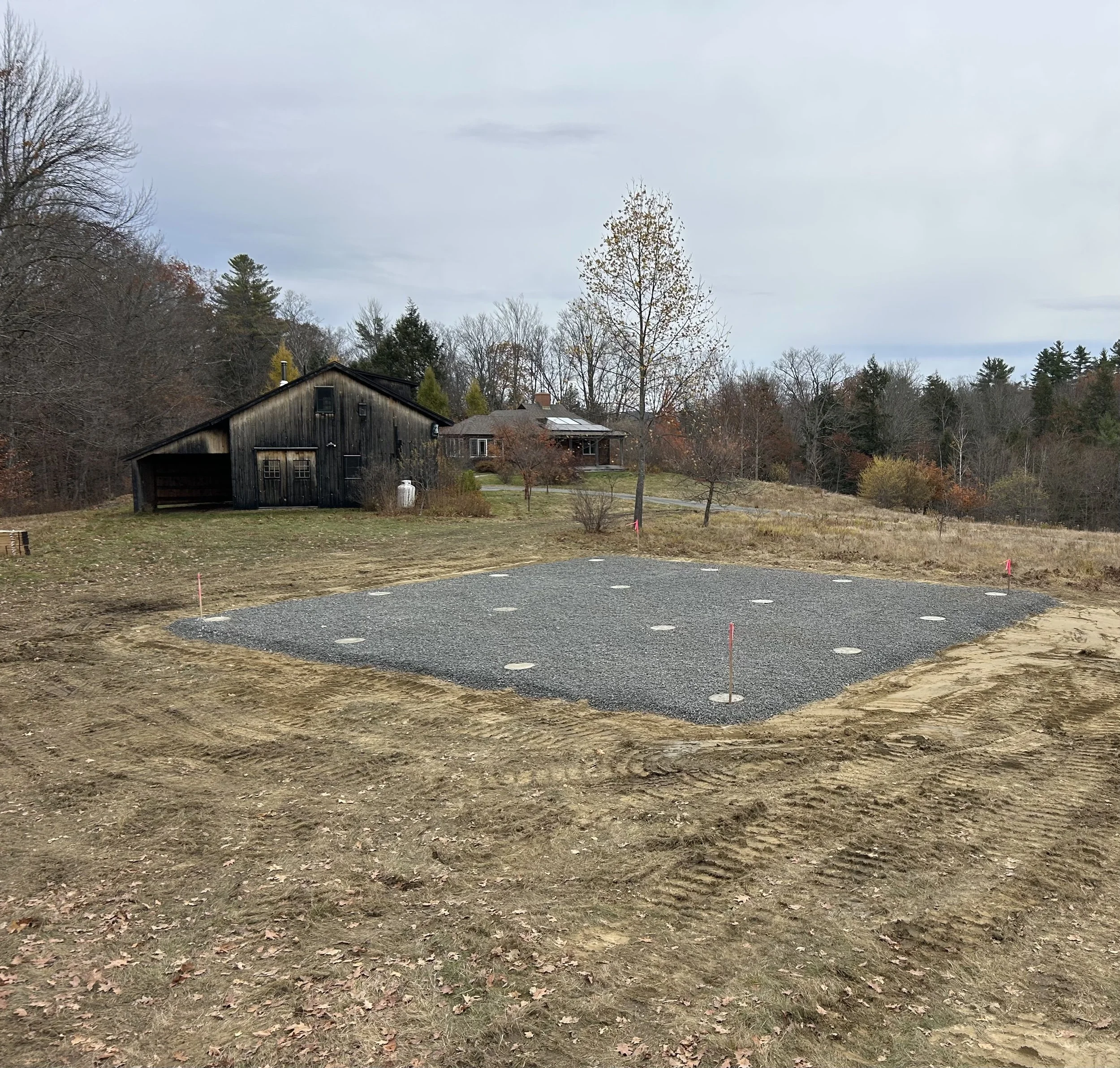 Construction site with a gravel foundation prepared for a building, in a rural area with trees and residential houses in the background.