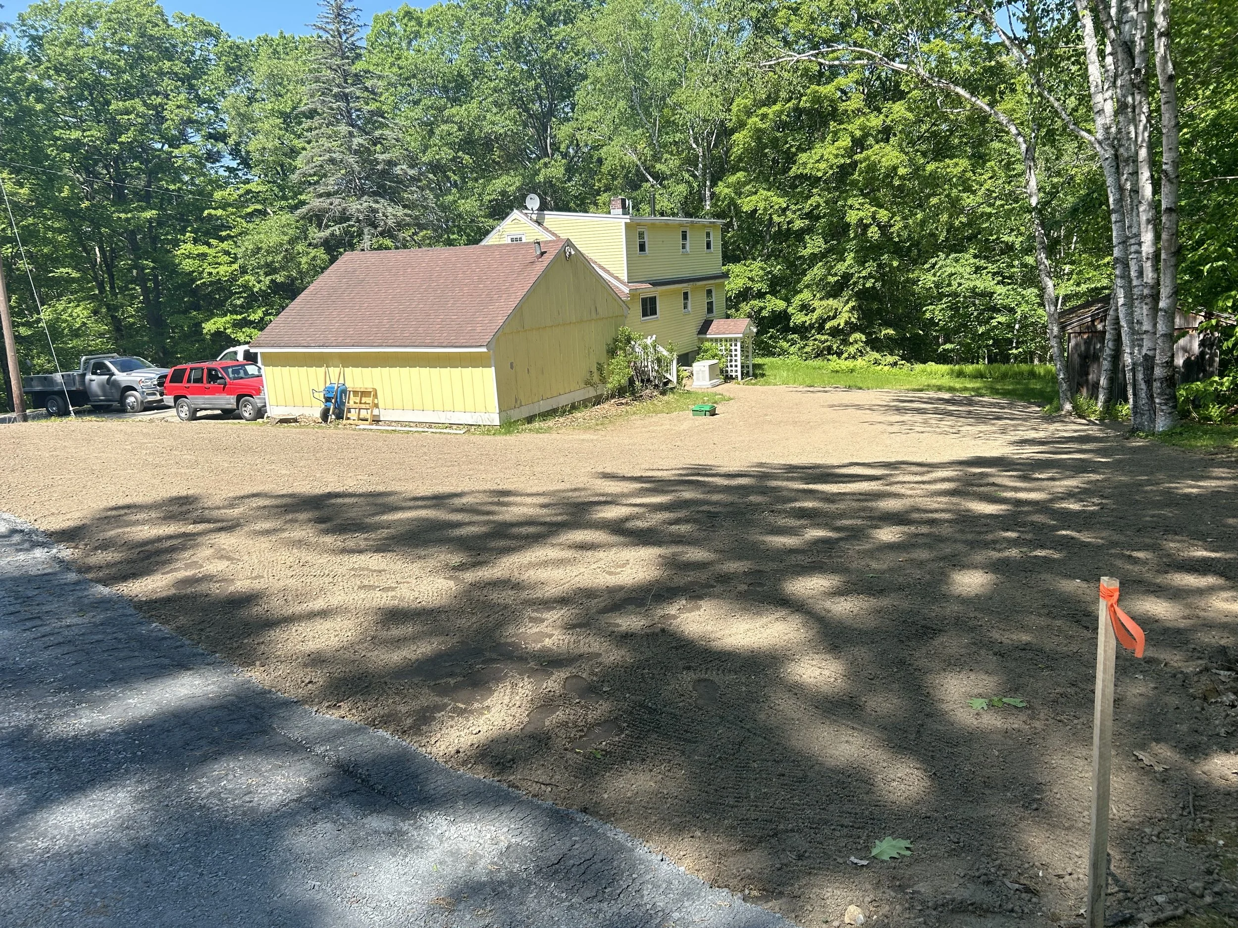 A cleared and leveled dirt lot in front of a yellow house with a brown roof, surrounded by green trees, with parked cars and construction stakes.