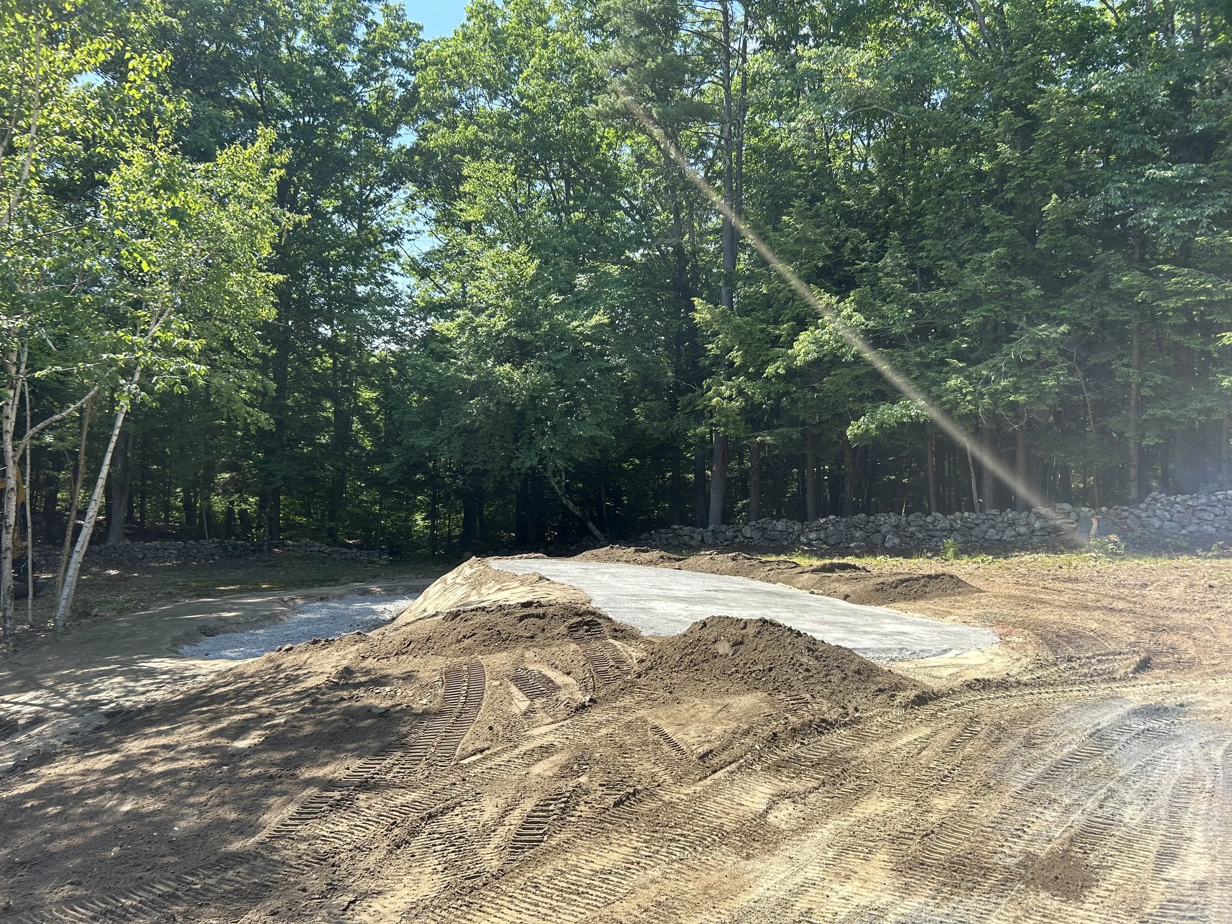 Construction site with a dirt road, some construction materials, and a wooded background.