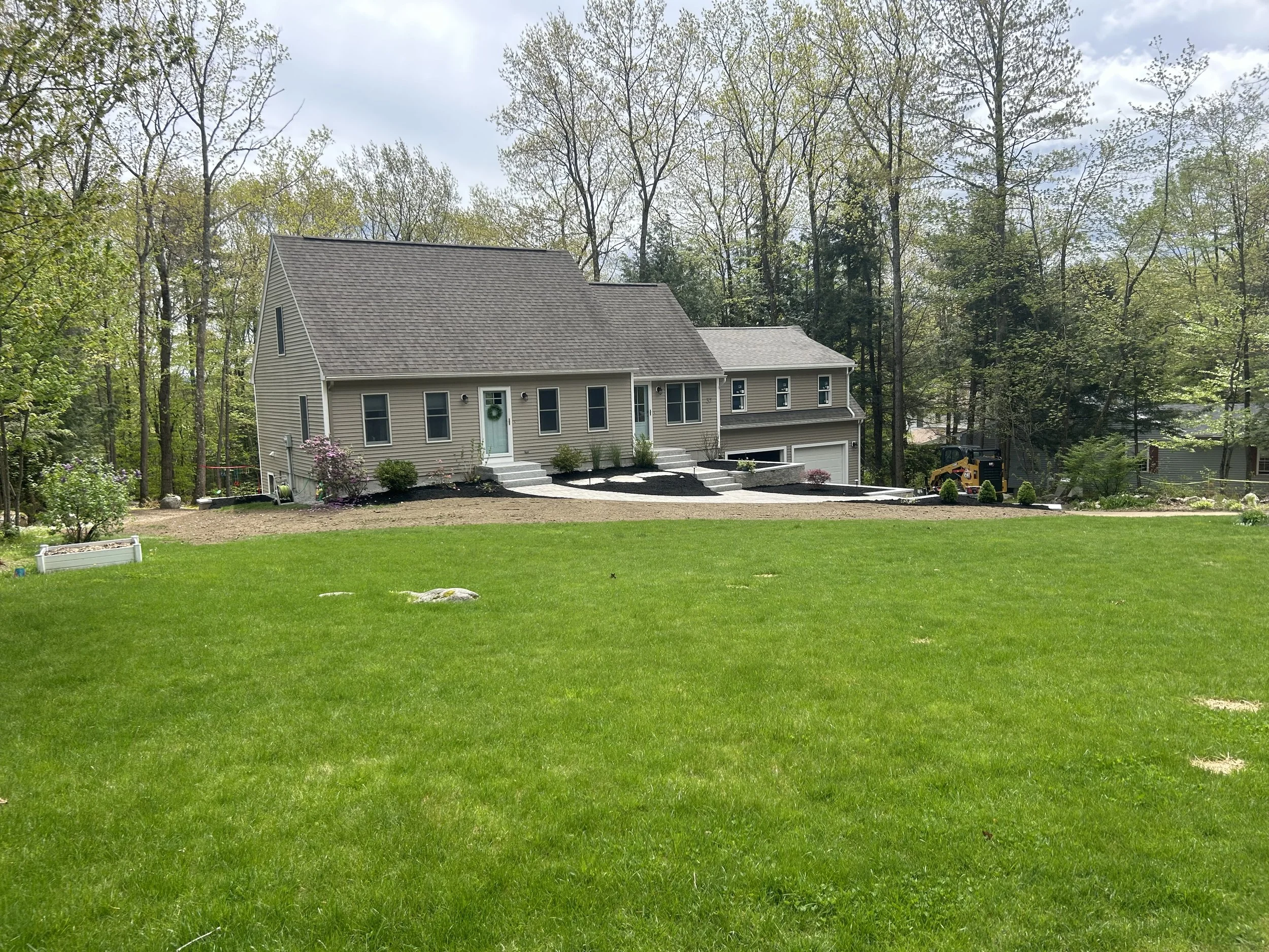 A house with a sloped roof situated at the edge of a wooded area, with well-maintained lawn and landscaping in the front, and a small construction vehicle parked near the back.