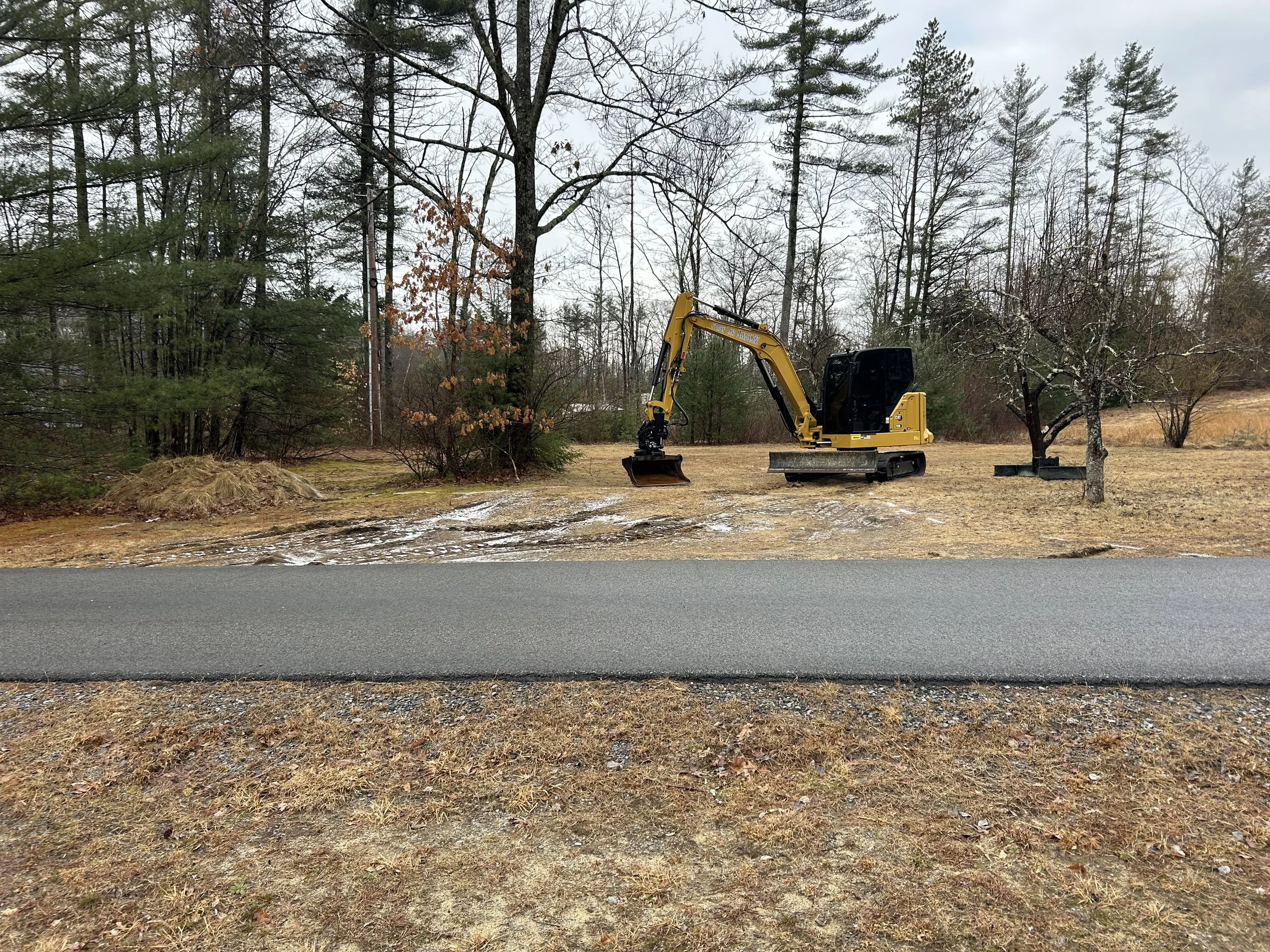 A yellow mini excavator working on a patch of land next to a paved road, surrounded by trees and a cloudy sky.