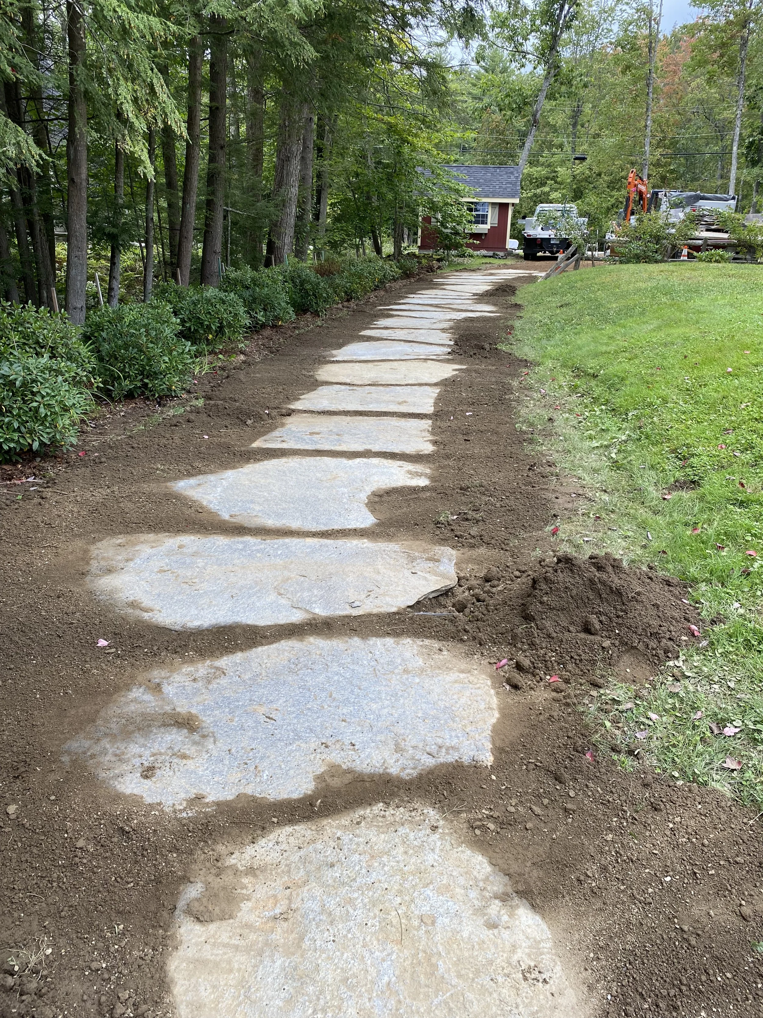 A freshly laid stone pathway winding through a wooded yard with green shrubs and a small red house in the background, some construction equipment and trucks visible.