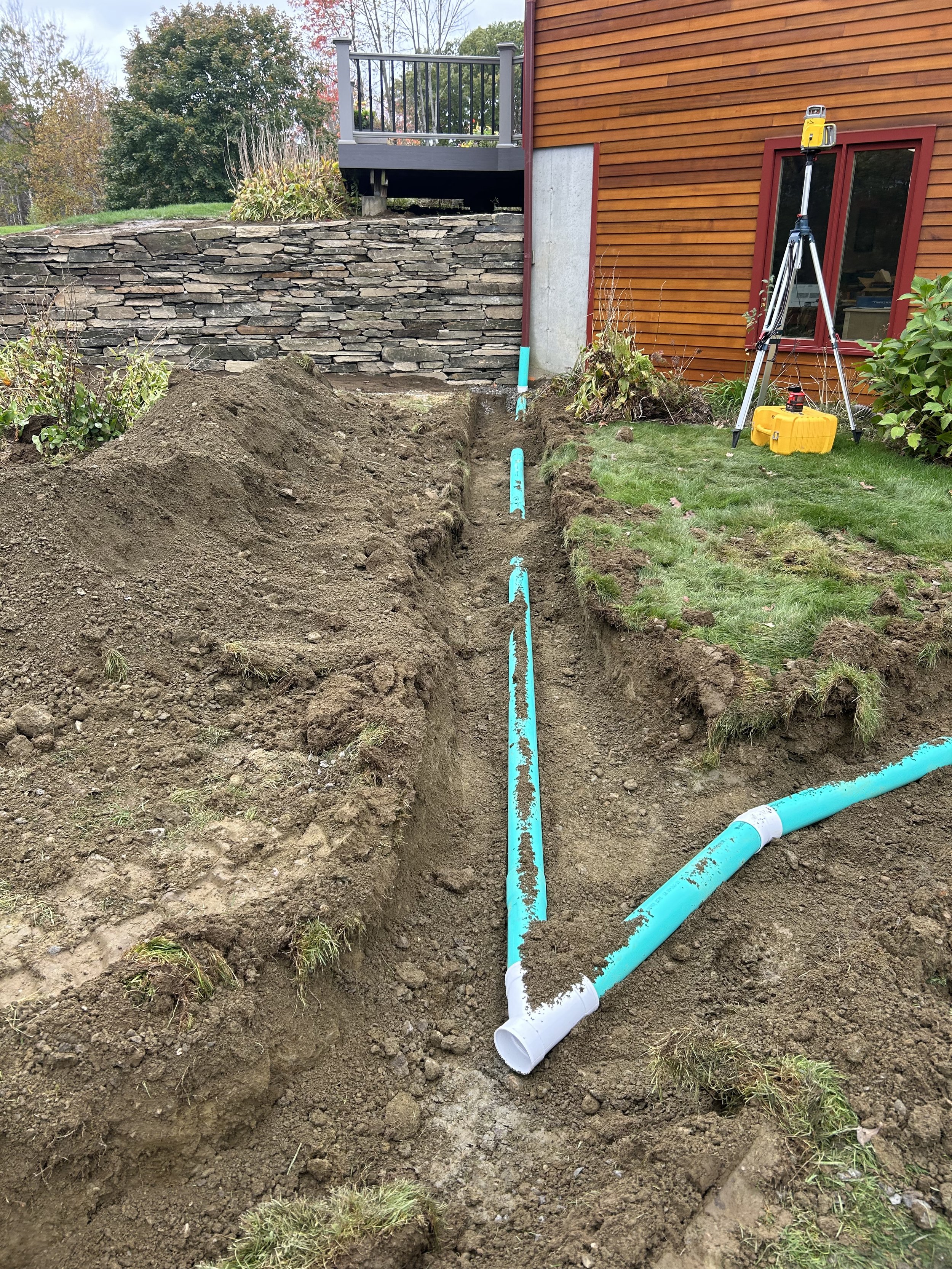 Installation of underground plumbing pipes in a backyard excavation with a trench, near a house with a wooden exterior, outdoor tools, and equipment.