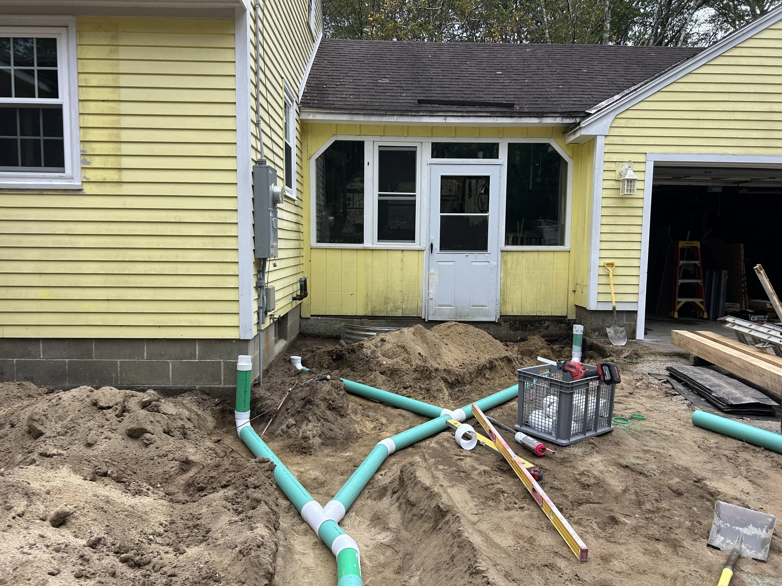 Construction site in backyard with dirt, pipes, tools, and a partially excavated area near a yellow house with a white door and garage.