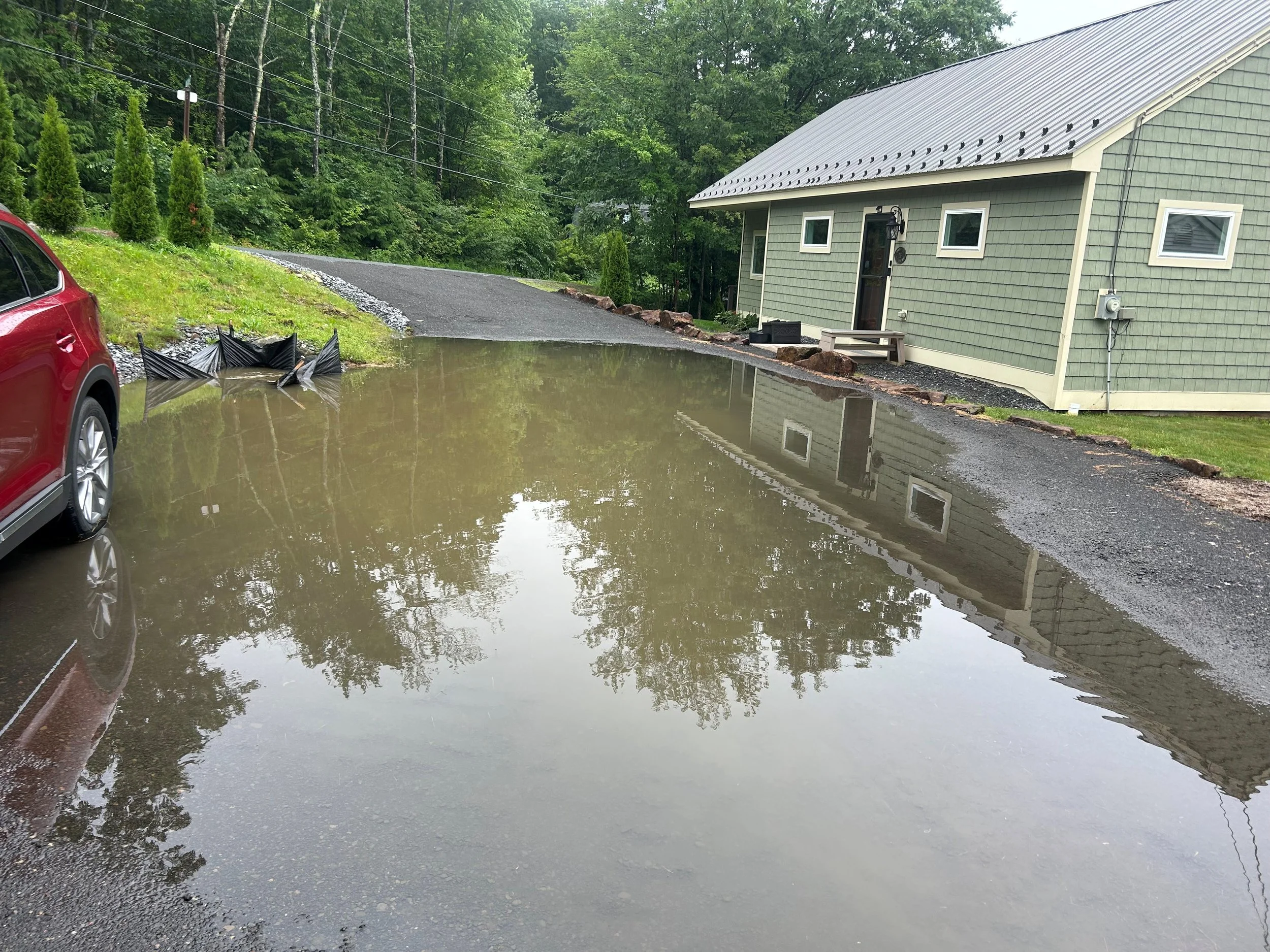 A flooded parking area with a large puddle covering the ground next to a green house and a red vehicle.