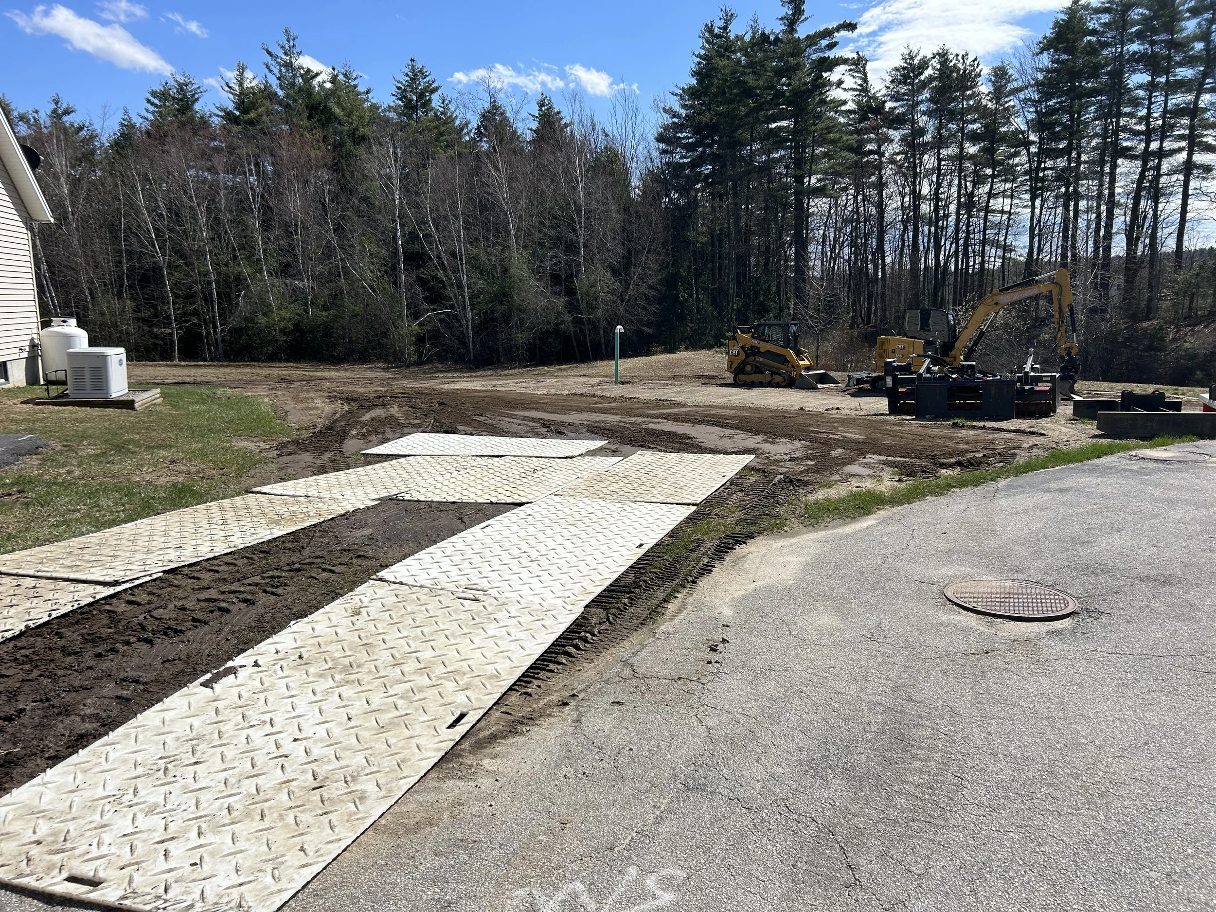 Construction site with dirt road, metal plates, and small yellow excavators near trees and a house.