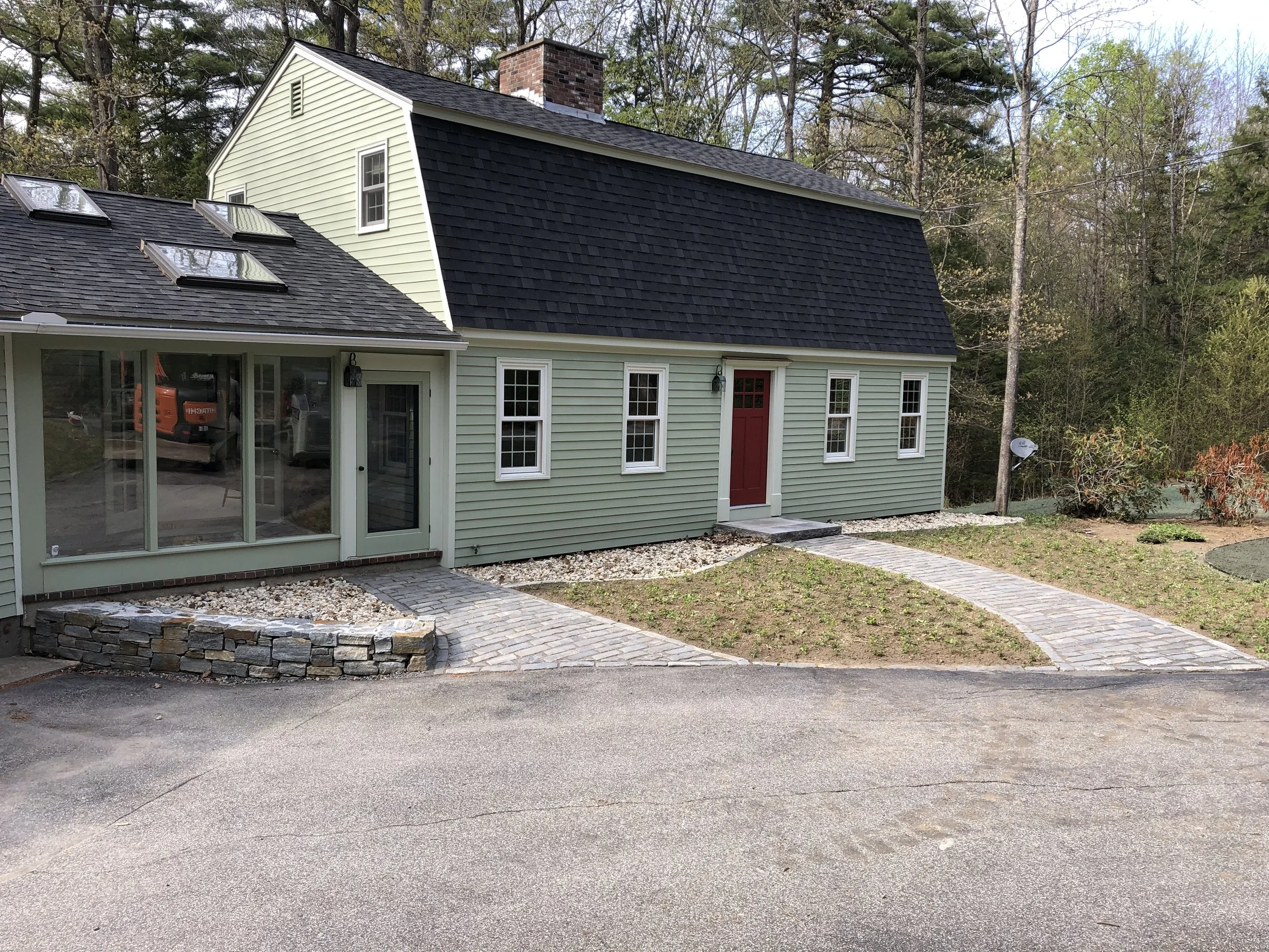 A house with a light green exterior, multiple windows, a red door, and a sloped roof with black shingles, surrounded by trees.