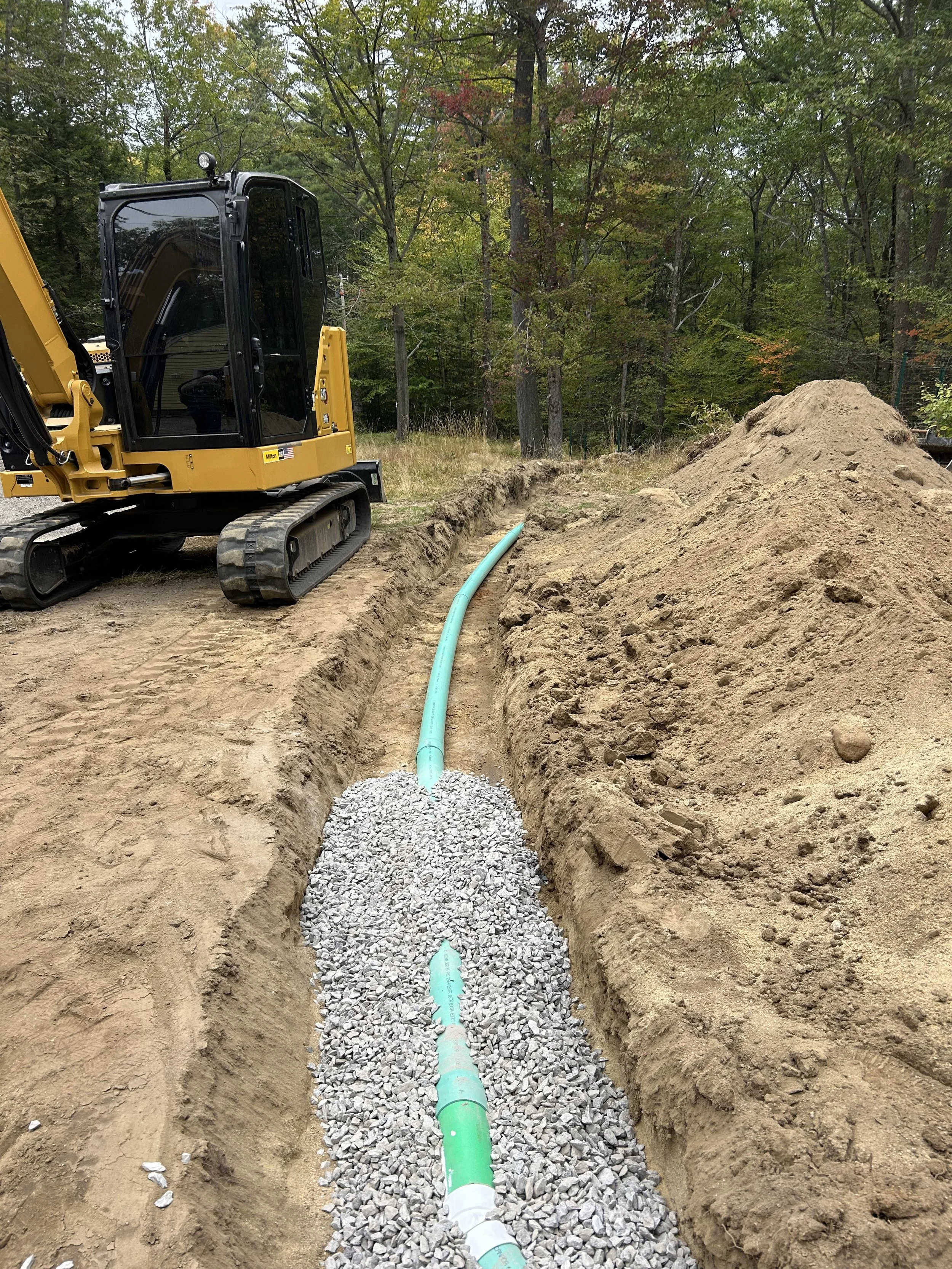 Construction site with a yellow excavator digging a trench for a water pipe system, surrounded by dirt and trees.