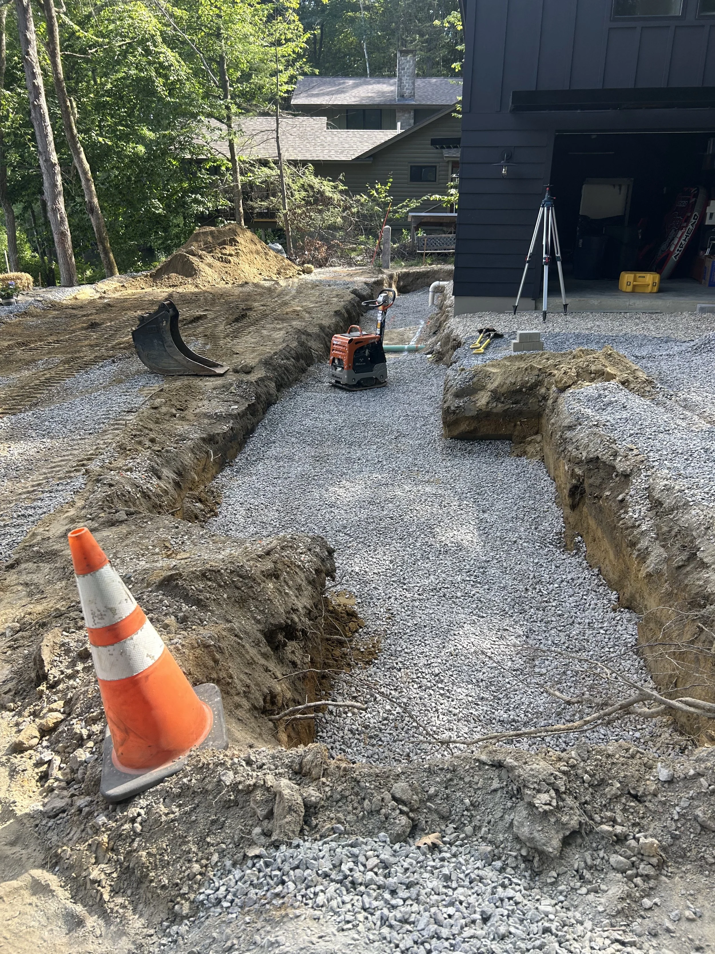 Construction site with a dug-out trench in front of a house, orange traffic cone in the foreground, gravel on the ground, construction tools, and equipment nearby.