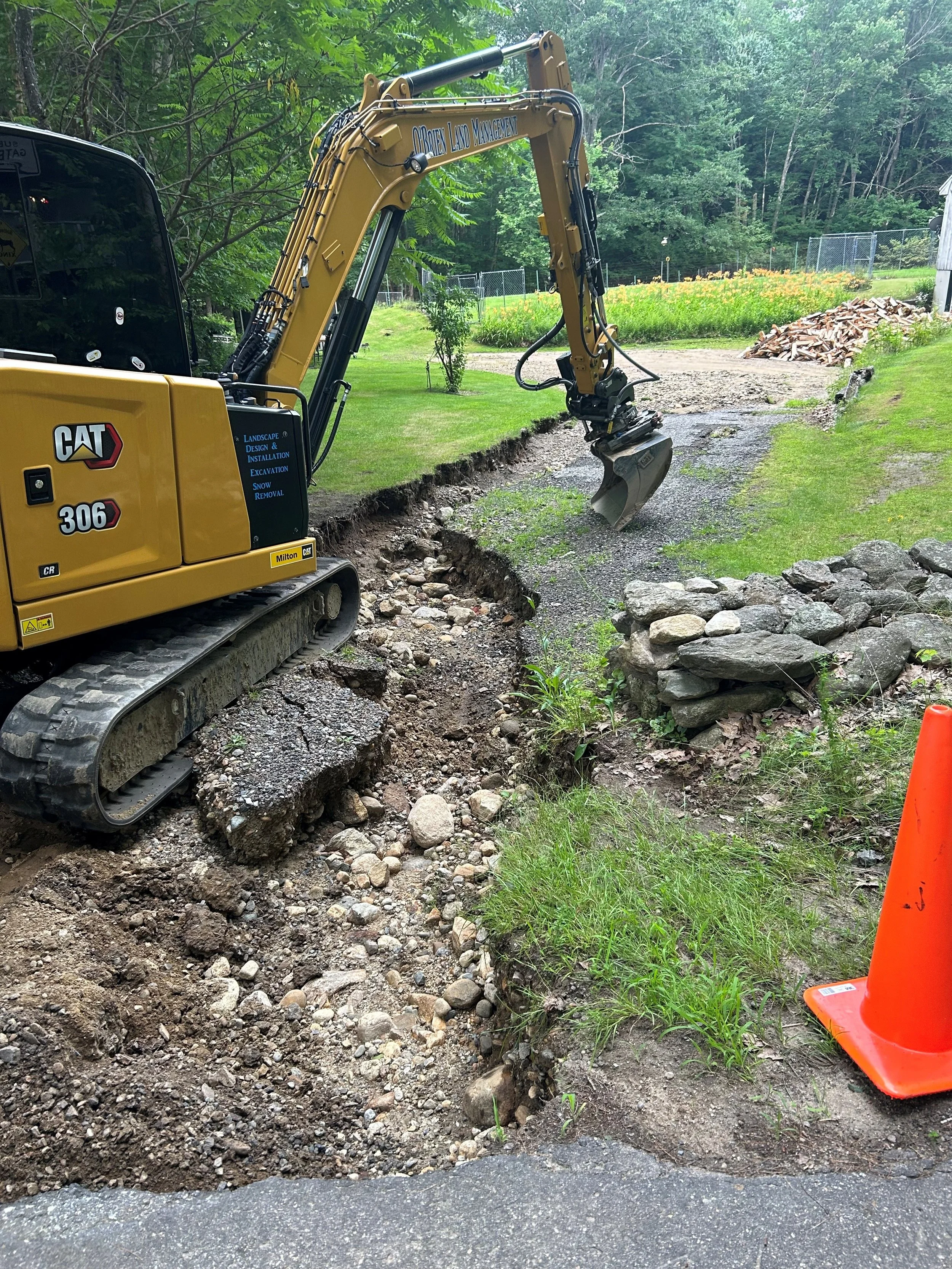 A yellow Caterpillar compact excavator digging a trench in a grassy area, with a pile of rocks to the side and an orange traffic cone nearby.