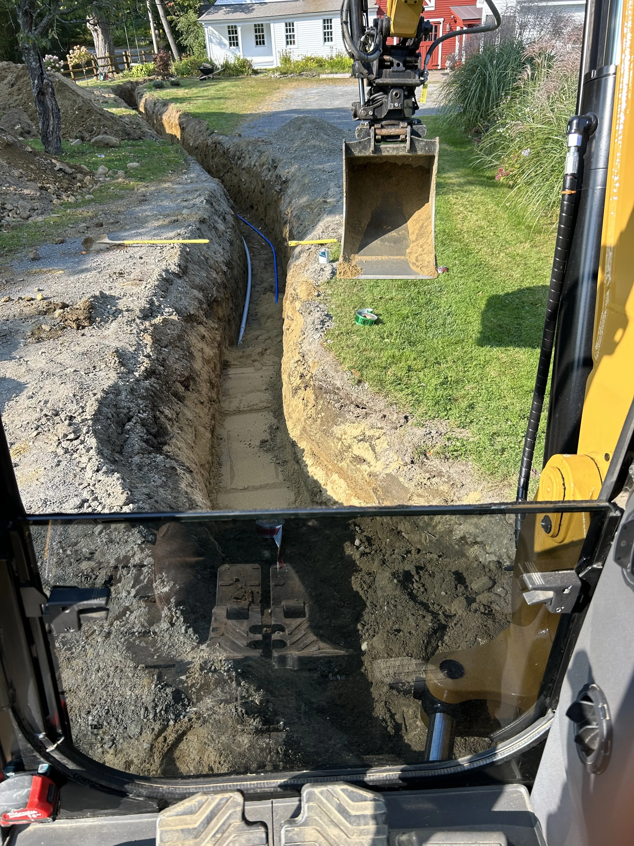 View from inside a construction vehicle looking out onto a trench being excavated for underground utility lines, with a house and garden in the background.