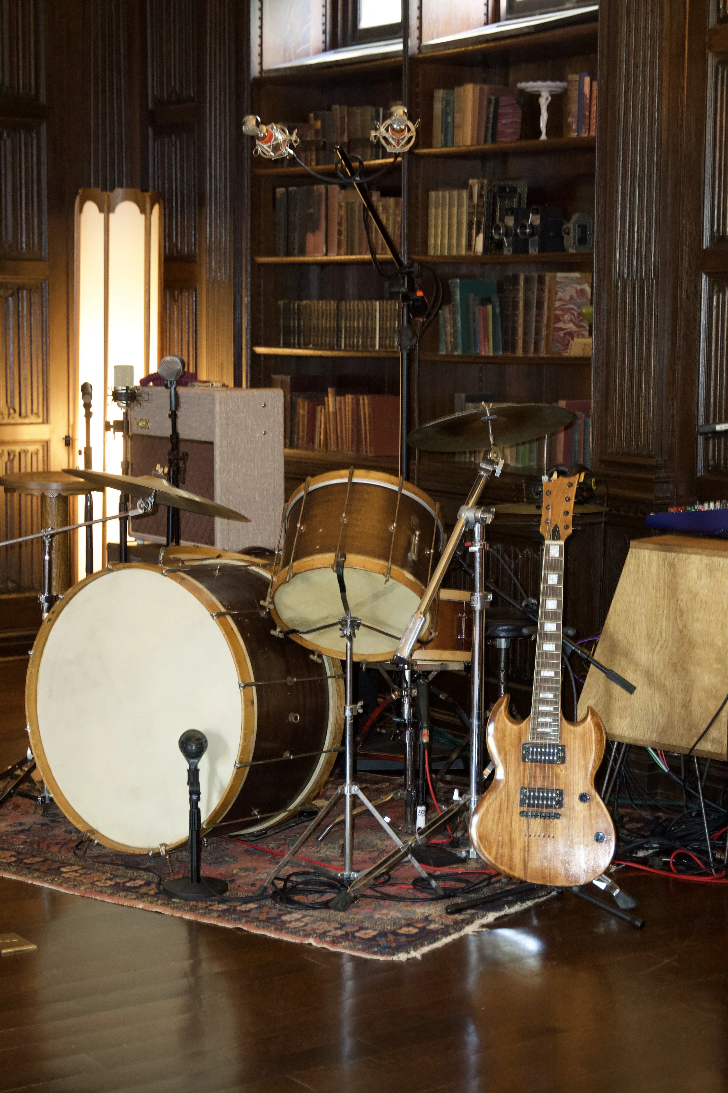 A music studio setup with a drum set, electric guitar, microphone, and amplifiers in a room with wooden walls and bookshelves.
