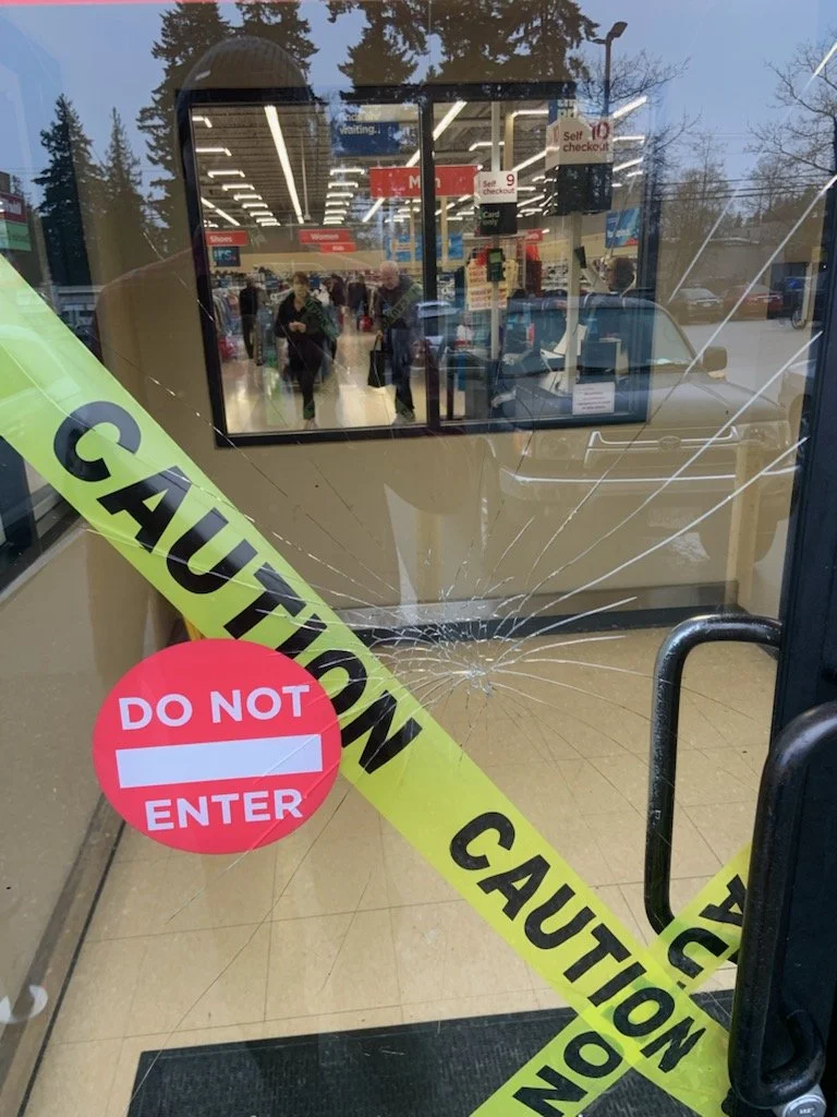Broken glass door with caution tape reading 'CAUTION' and a red circle sign saying 'Do Not Enter'. The interior of a store with shoppers visible through the glass, and reflections of trees and parked cars outside.
