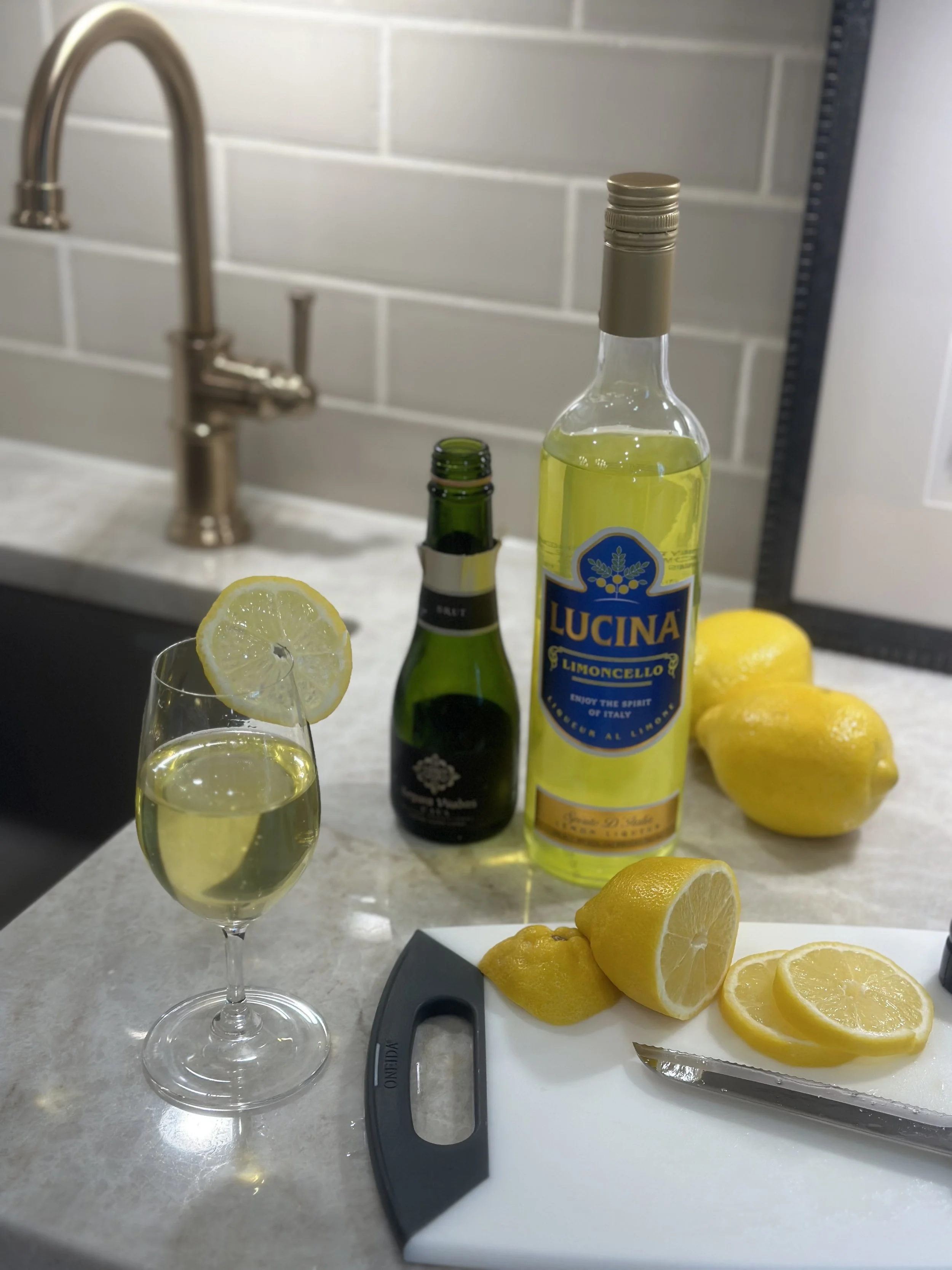 A glass of lemon flavored alcohol with lemon slice, a bottle of Lucina Limoncello, a bottle of champagne, and fresh lemons on a kitchen counter.