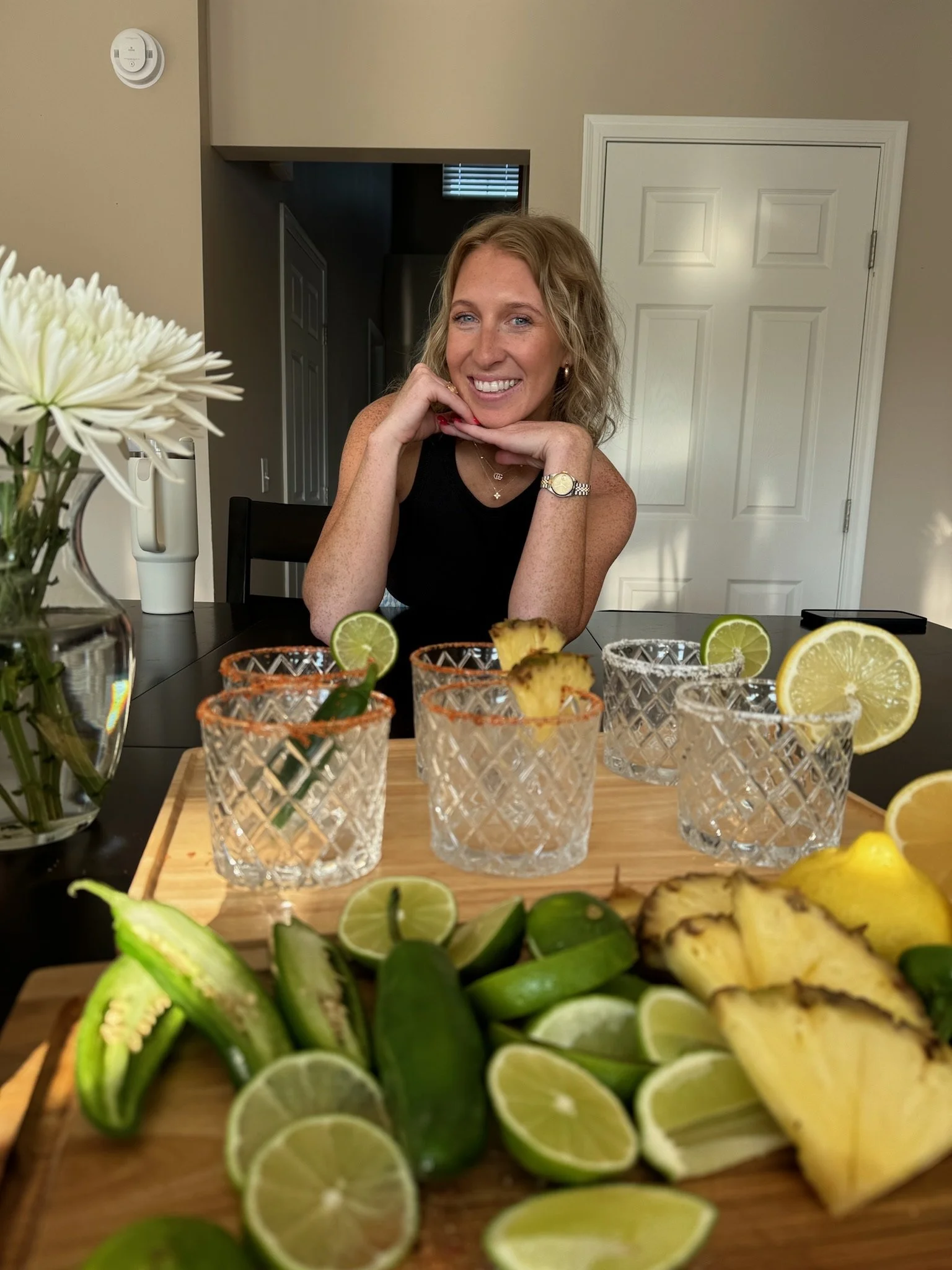 Barmaid Emma smiling at a table with six glass cups garnished with lime slices, and a wooden board with sliced limes, pineapples, and cucumbers. There is a white flower in a vase on the left side of the table.