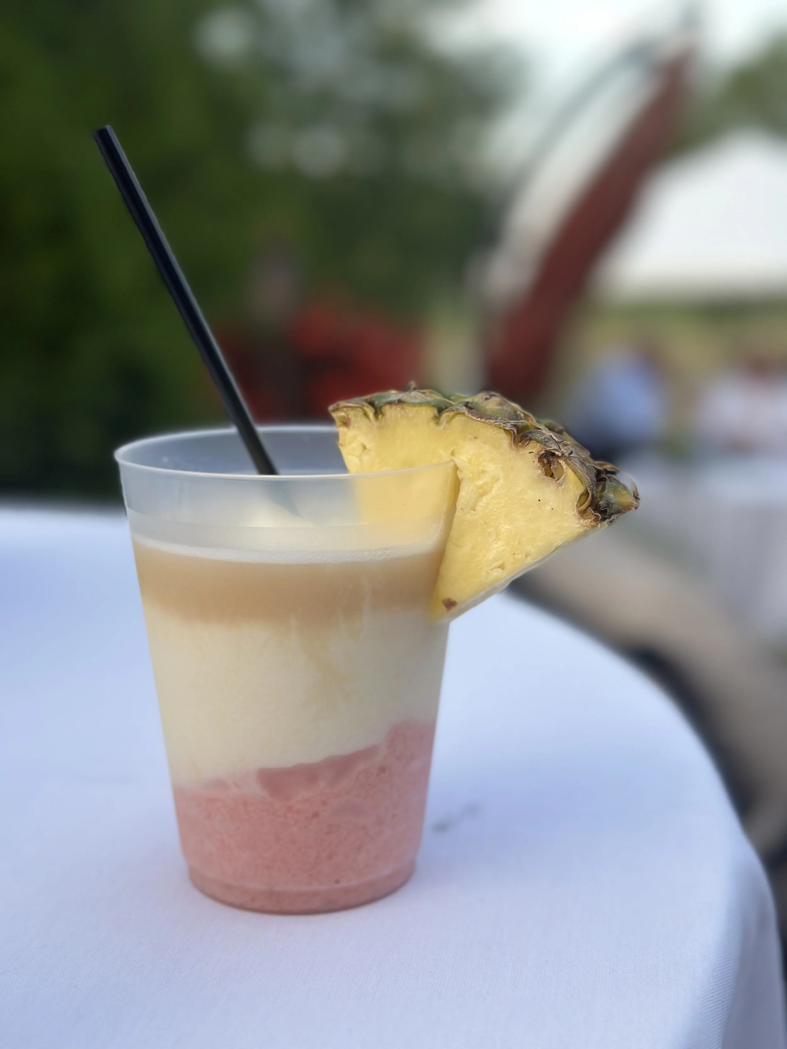 A layered fruit cocktail in a clear plastic cup with a pineapple wedge garnish and a black straw, set on a white tablecloth with an outdoor background.