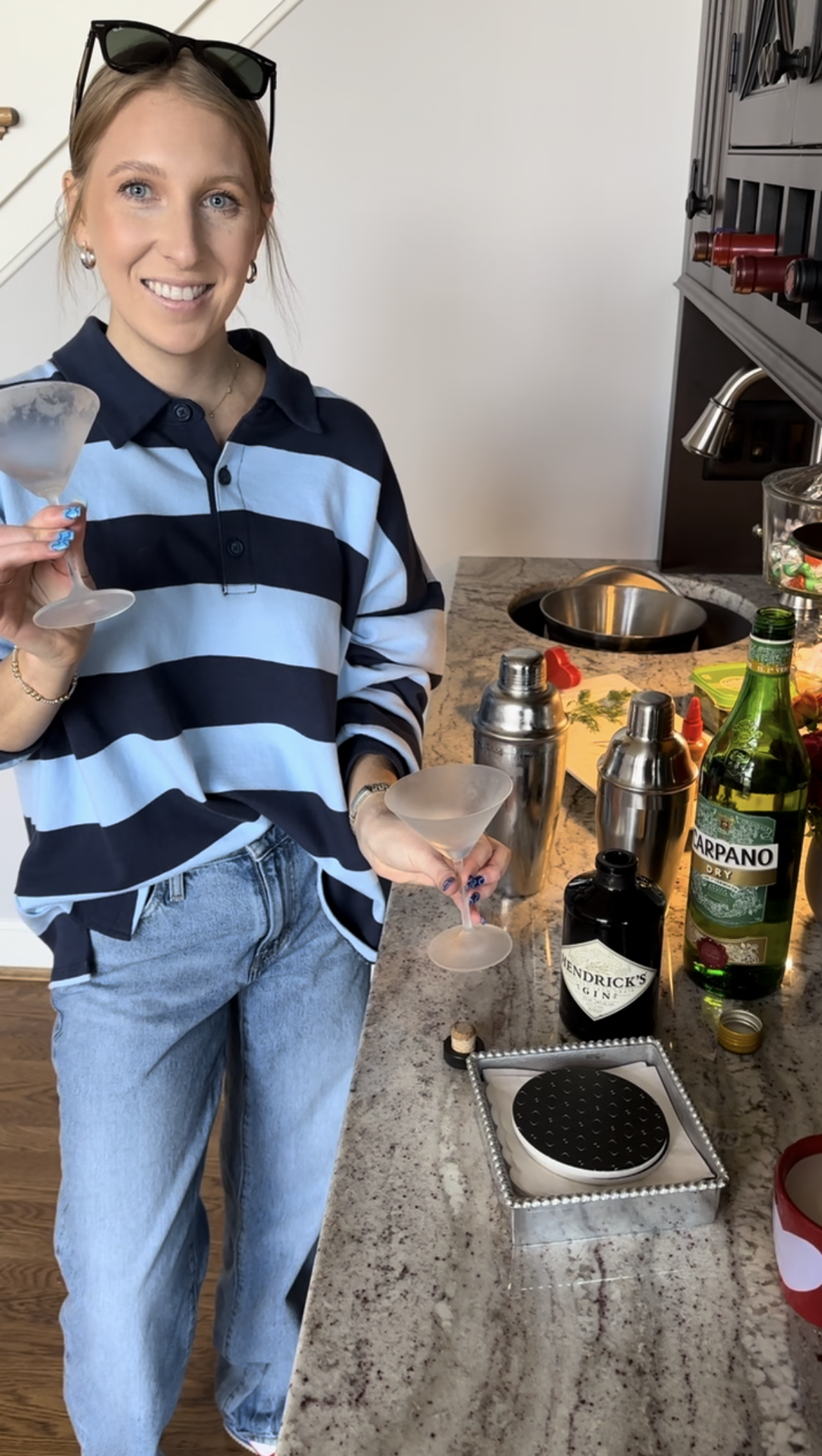 A woman smiling and holding two cocktail glasses in a kitchen. The countertop has bartender tools, a bottle of Carpano dry vermouth, Hendrick's gin, and a shaker. The woman is wearing a blue and black striped polo shirt, light-wash jeans, sunglasses on her head, and pearl earrings.