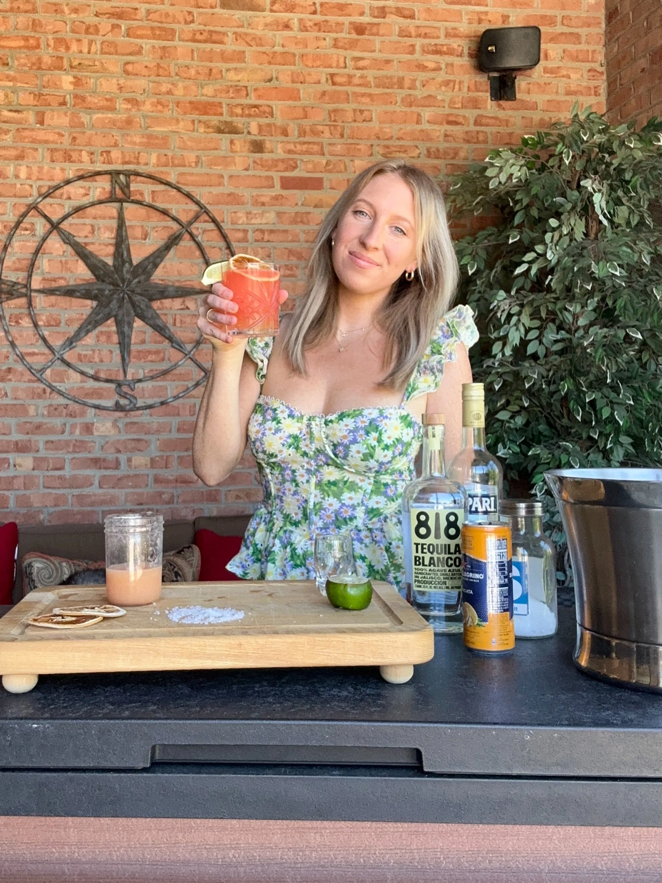 A woman standing behind a bar counter holding a colorful cocktail garnished with a lime wedge. The bar has bottles of alcohol, a lime, salt, and other ingredients on a wooden paddle. The background features a brick wall, a decorative metal compass rose, and a green leafy plant.