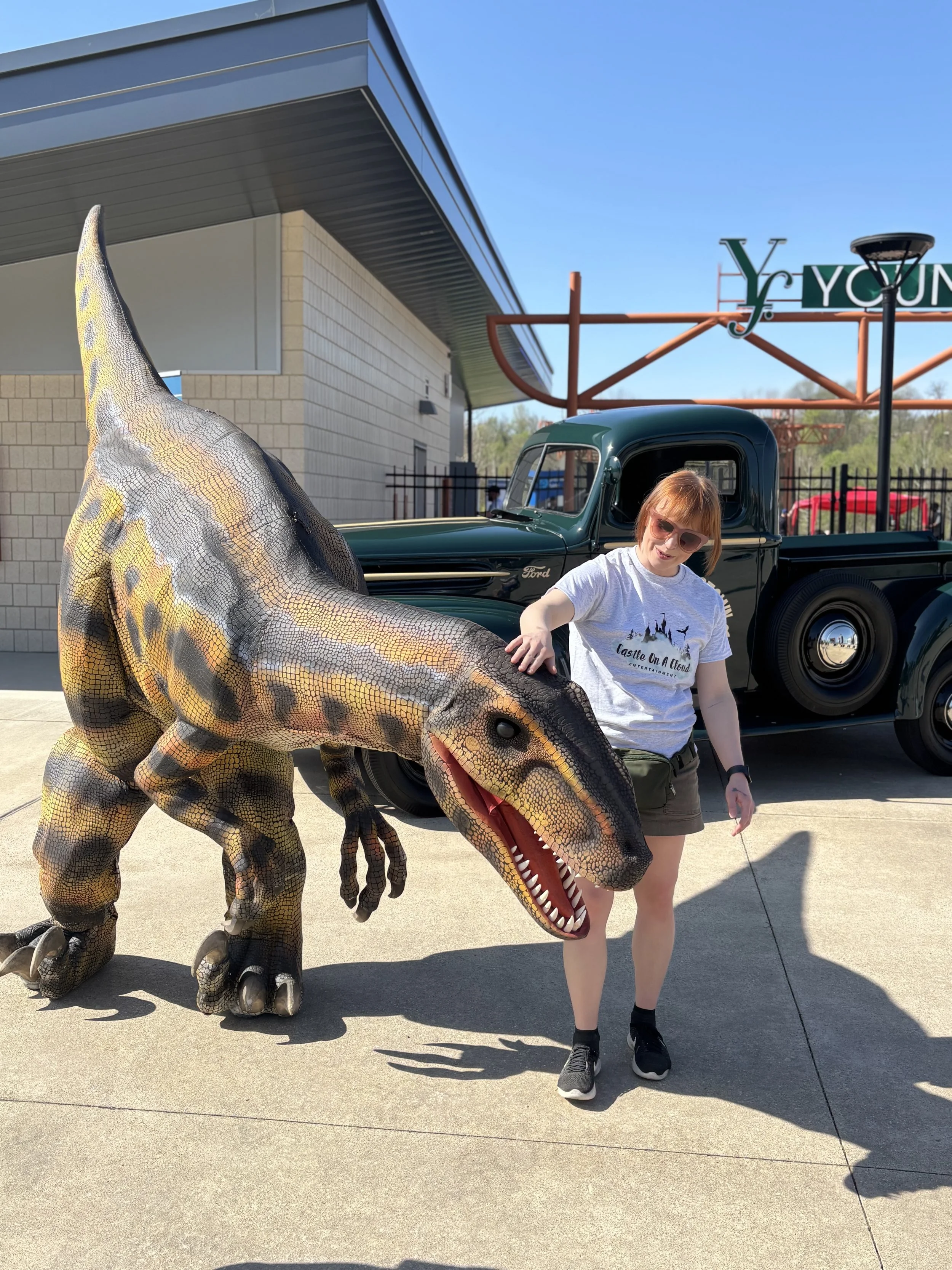 A girl with red hair and sunglasses standing next to a realistic-looking dinosaur costume resembling a T-Rex, in front of a vintage green Ford truck and an old-fashioned sign.