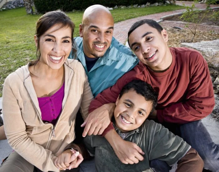 Una sonriente familia de cuatro sentados al aire libre en un banco del parque, incluyendo una mujer, un hombre, un adolescente y un niño pequeño, disfrutando de un día soleado con vegetación alrededor.