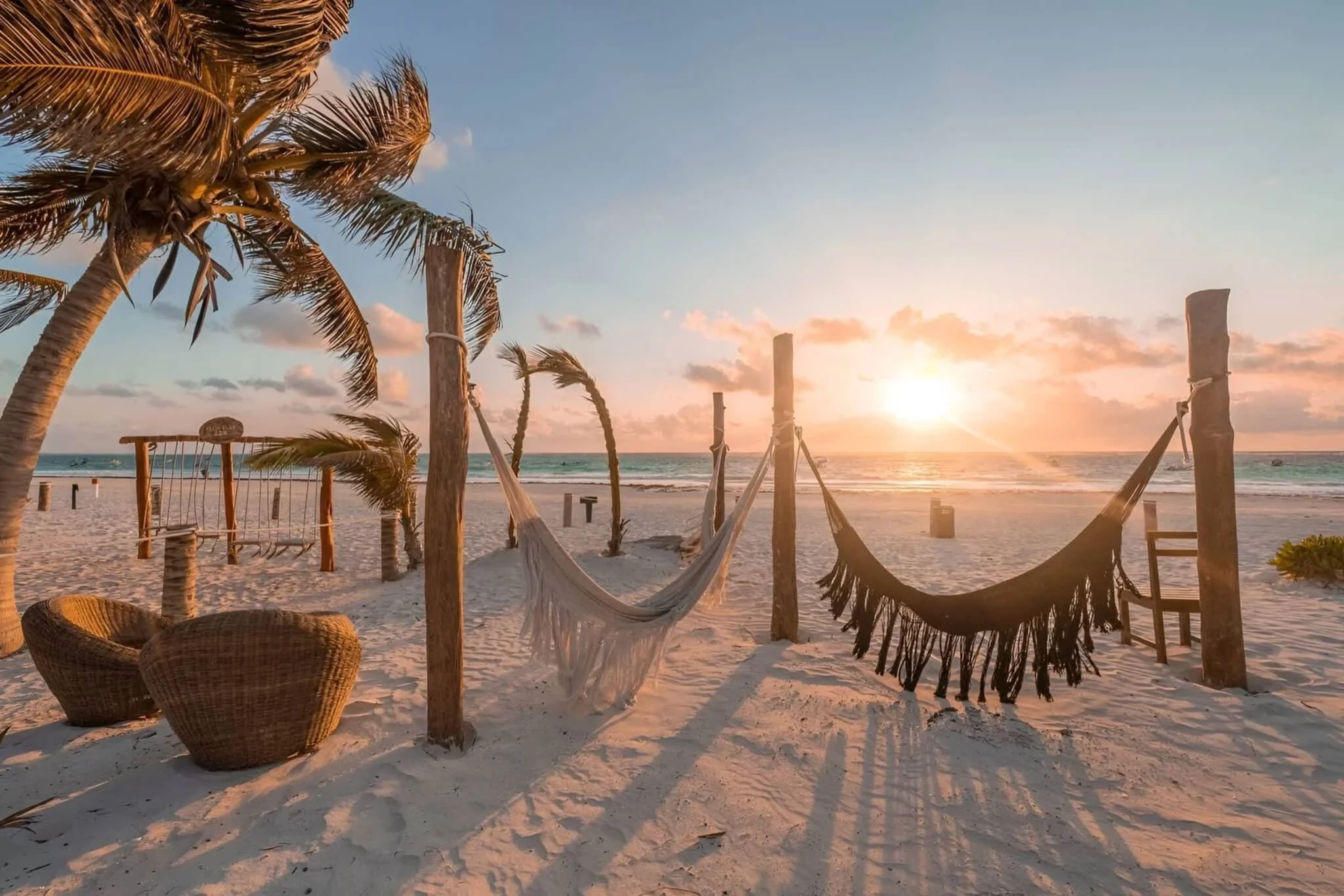 Escena de playa con palmeras, hamacas, sillas de mimbre y columpios al atardecer.