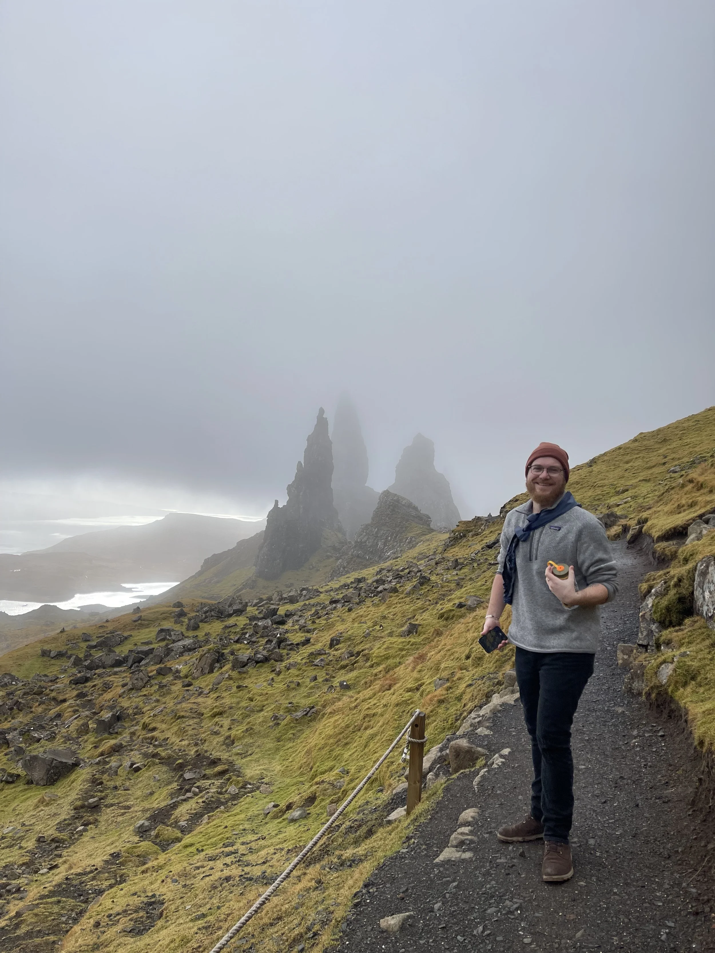 A man standing on a trail with rocky and grassy terrain, holding a phone and a yellow object, smiling, with misty mountains and rock formations in the background.