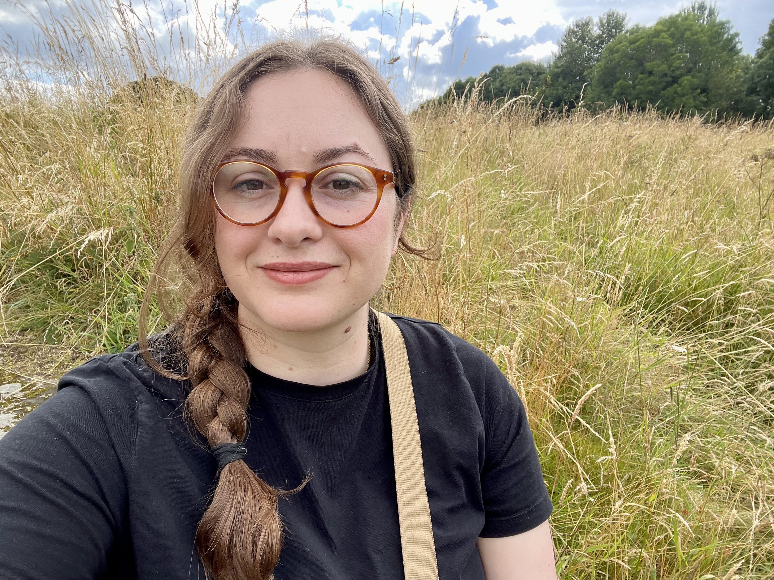 A woman with long brown hair in a braid, wearing glasses and a black t-shirt, sitting in a grassy field with tall, golden grass, under a partly cloudy sky.